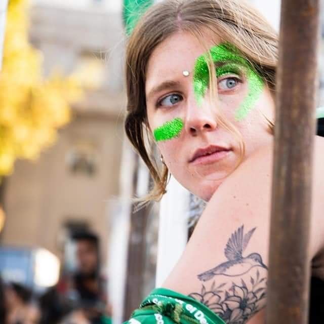 Närbild på en person med grön ansiktsmålning och en fågeltatuering på armen.