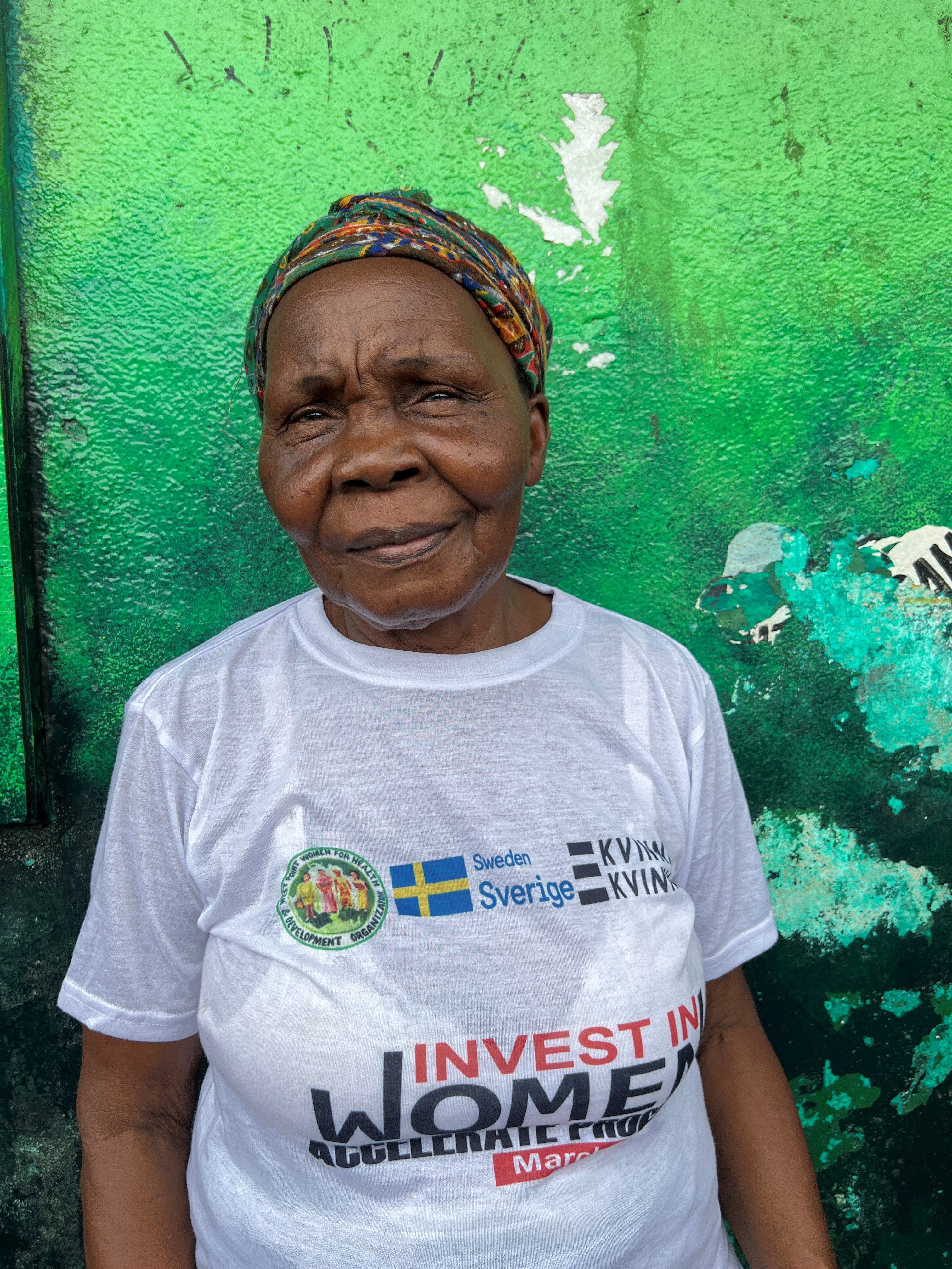 An older woman wearing a colorful headwrap looks toward the camera with a gentle smile against a green wall. She is wearing a white t-shirt printed with logos, including the Swedish flag, and the text "INVEST IN WOMEN".