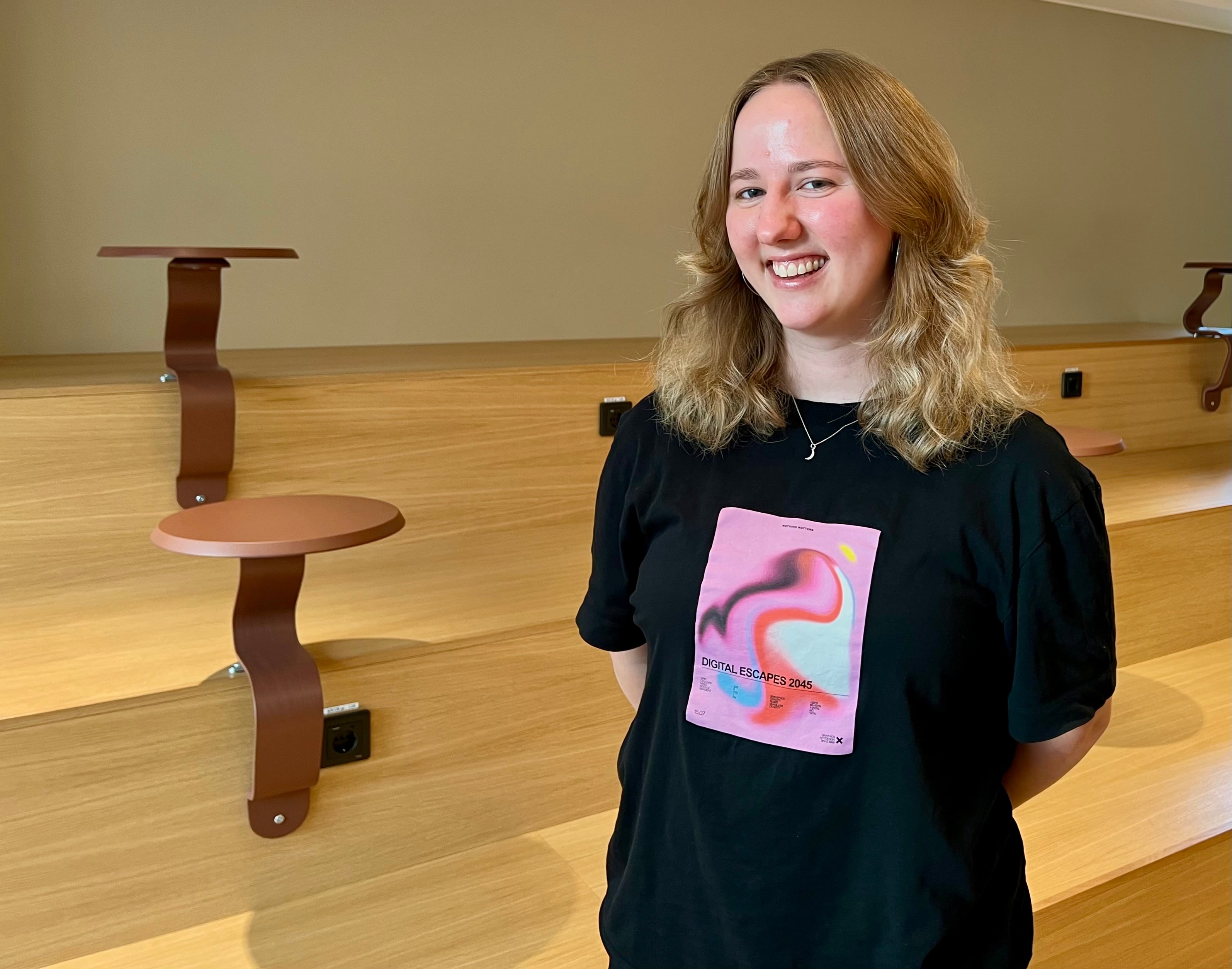 Leende person i svart t-shirt med ett rosa motiv framför en trätrappa.