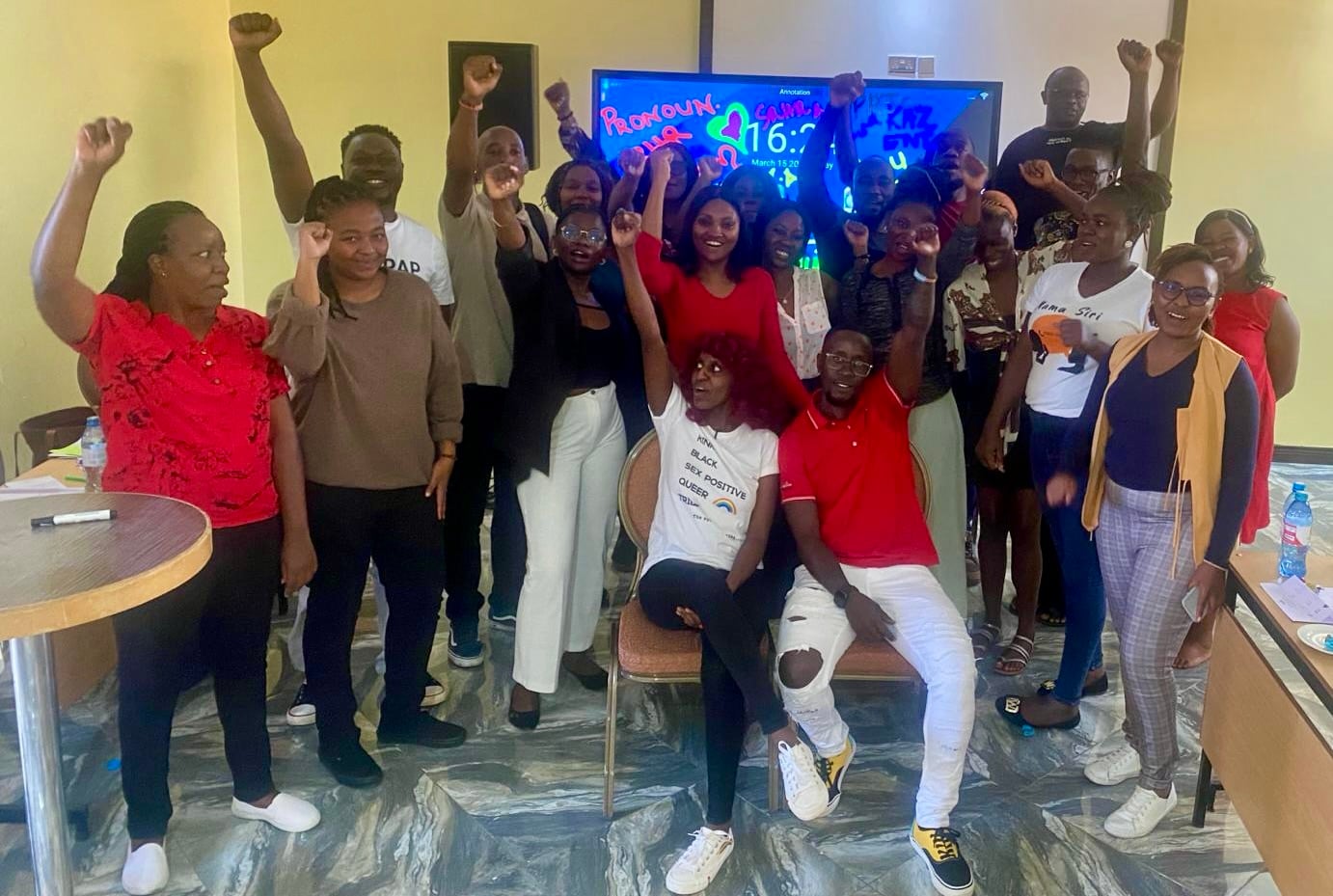 A group of people pose together indoors with their fists triumphantly raised in the air. They stand in a bright room with a polished, marbled floor in front of a large digital screen.