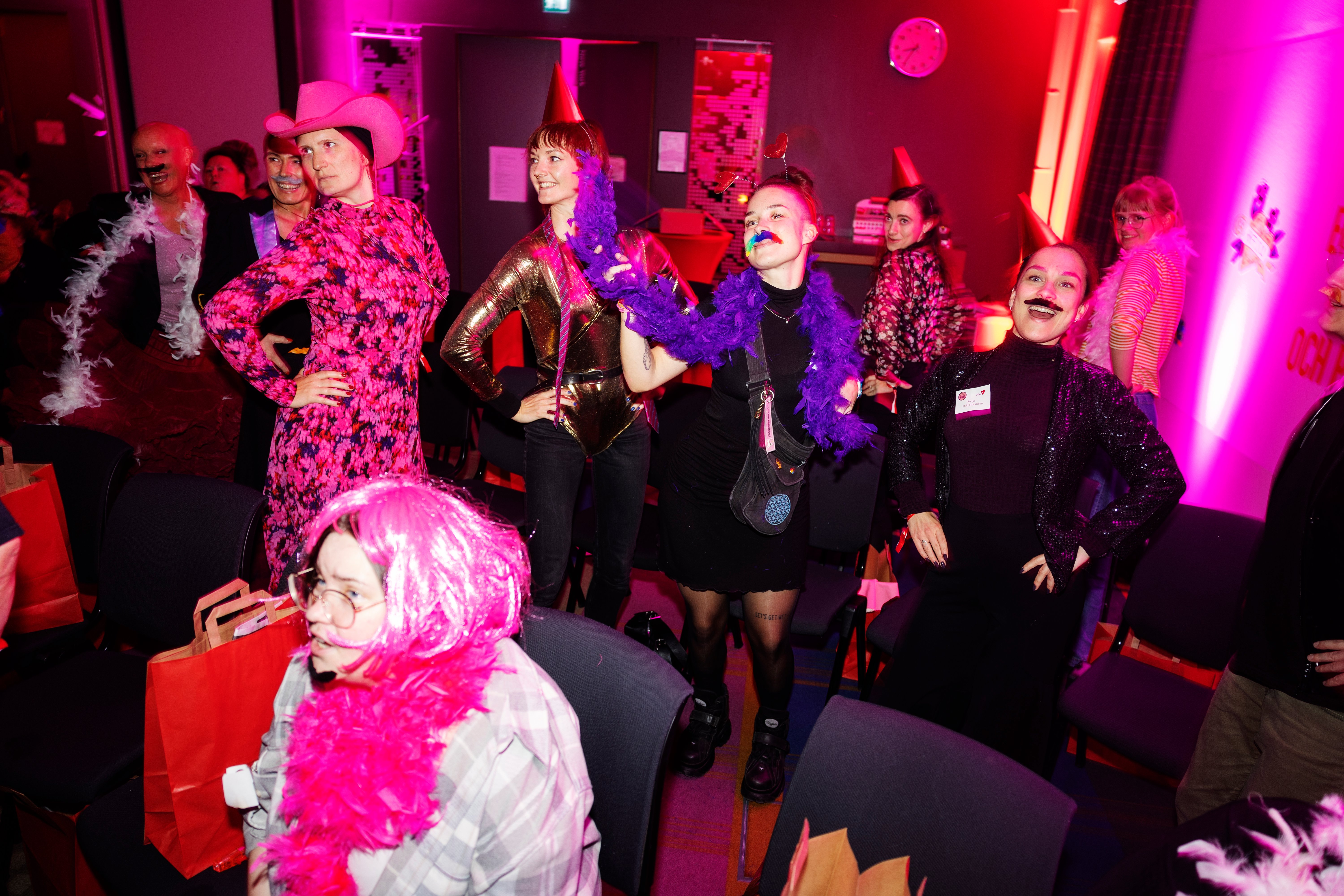 En grupp deltagare i färgglada peruker, boaormar och hattar dansar och poserar under en festkväll.