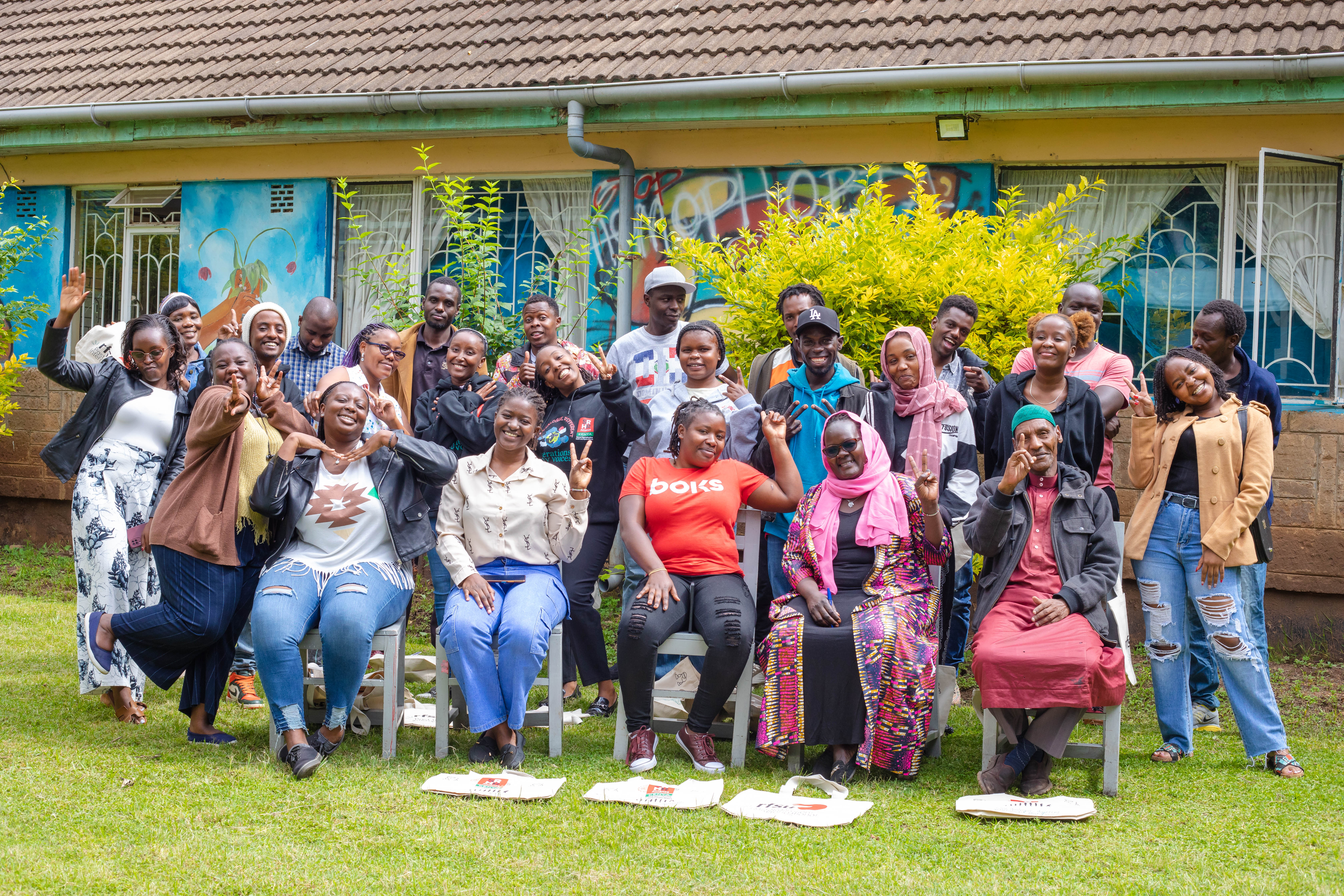 A large group of people smile and make hand gestures while posing for a photo on a green lawn outside a building. Several individuals are seated in the front row, with white tote bags resting on the grass in front of them.