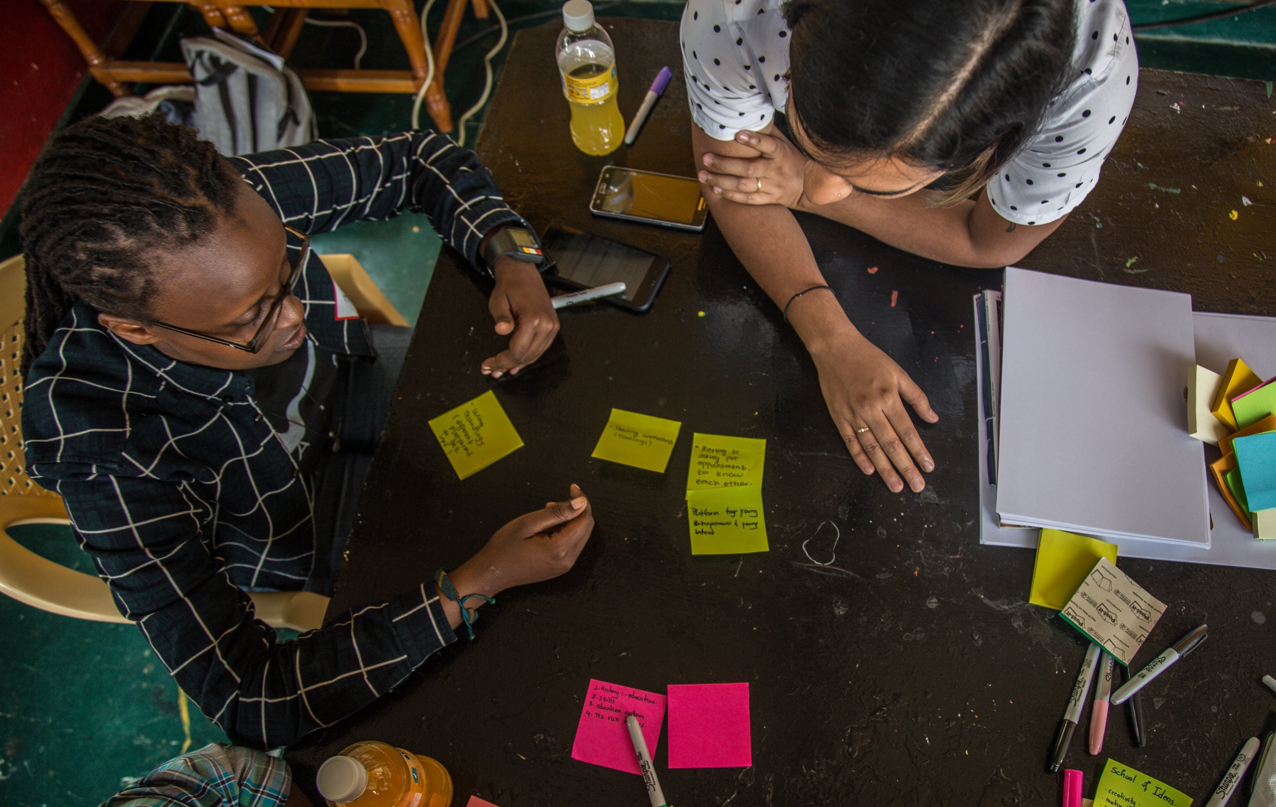 An overhead view shows two women collaborating around a dark table filled with handwritten sticky notes, notebooks, and pens during a brainstorming session. One woman wears glasses and a plaid shirt, while the other leans over the table in a polka-dot top.