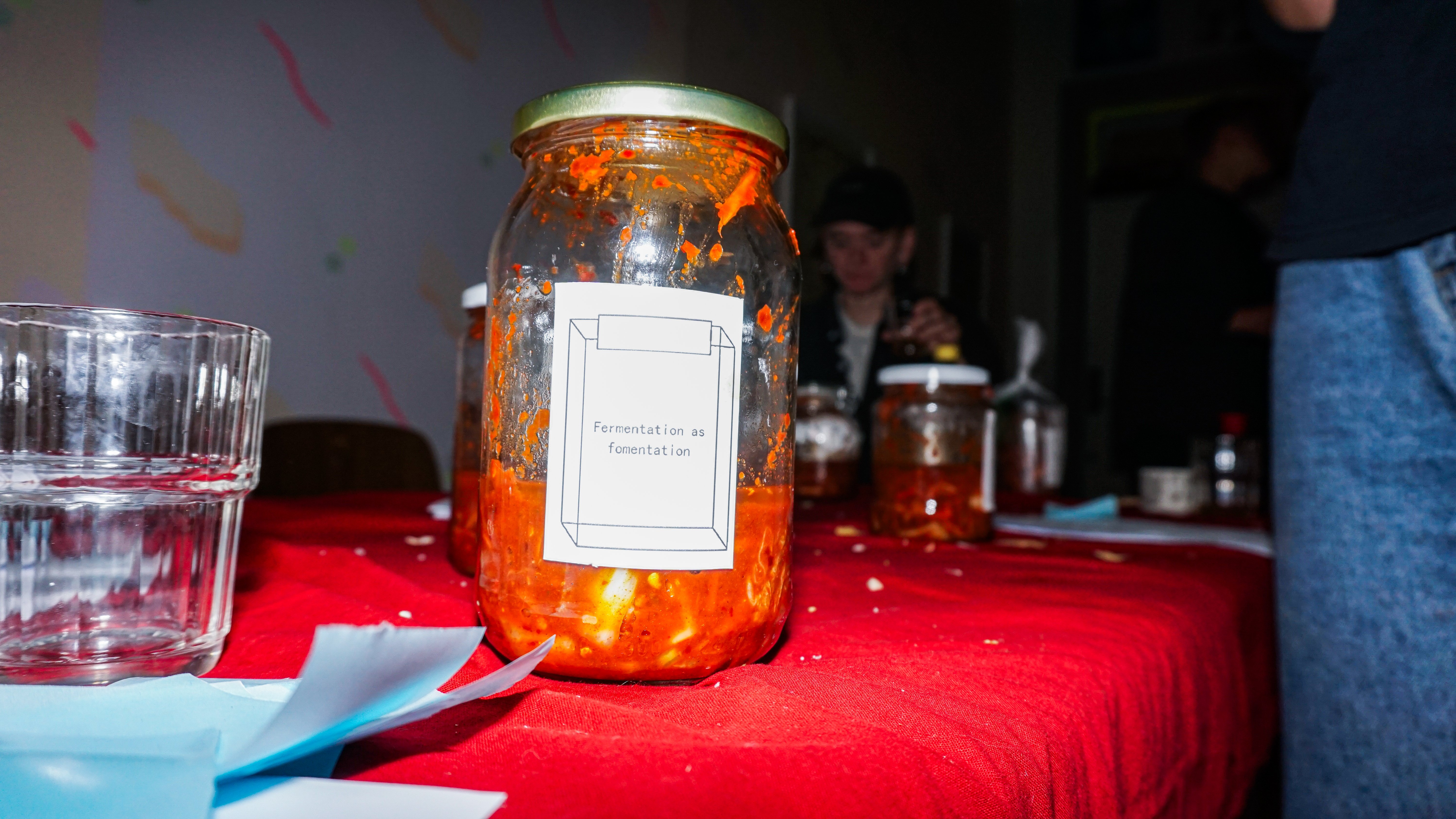 En glasburk med rött innehåll står på en röd duk med en etikett där det står "Fermentation as fomentation".