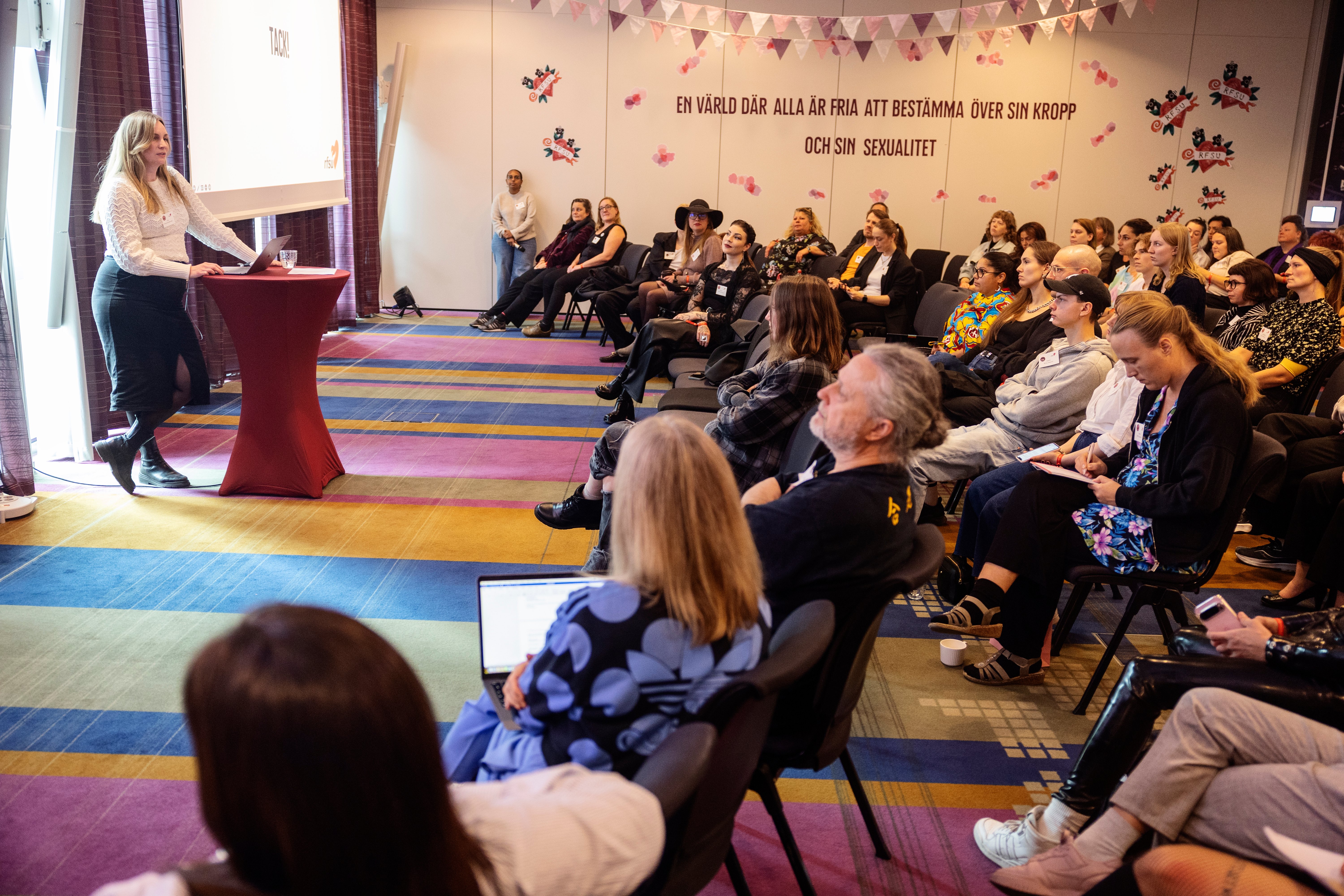 En kvinna presenterar från en talarstol för en stor grupp medlemmar som sitter i rader i en konferenslokal.