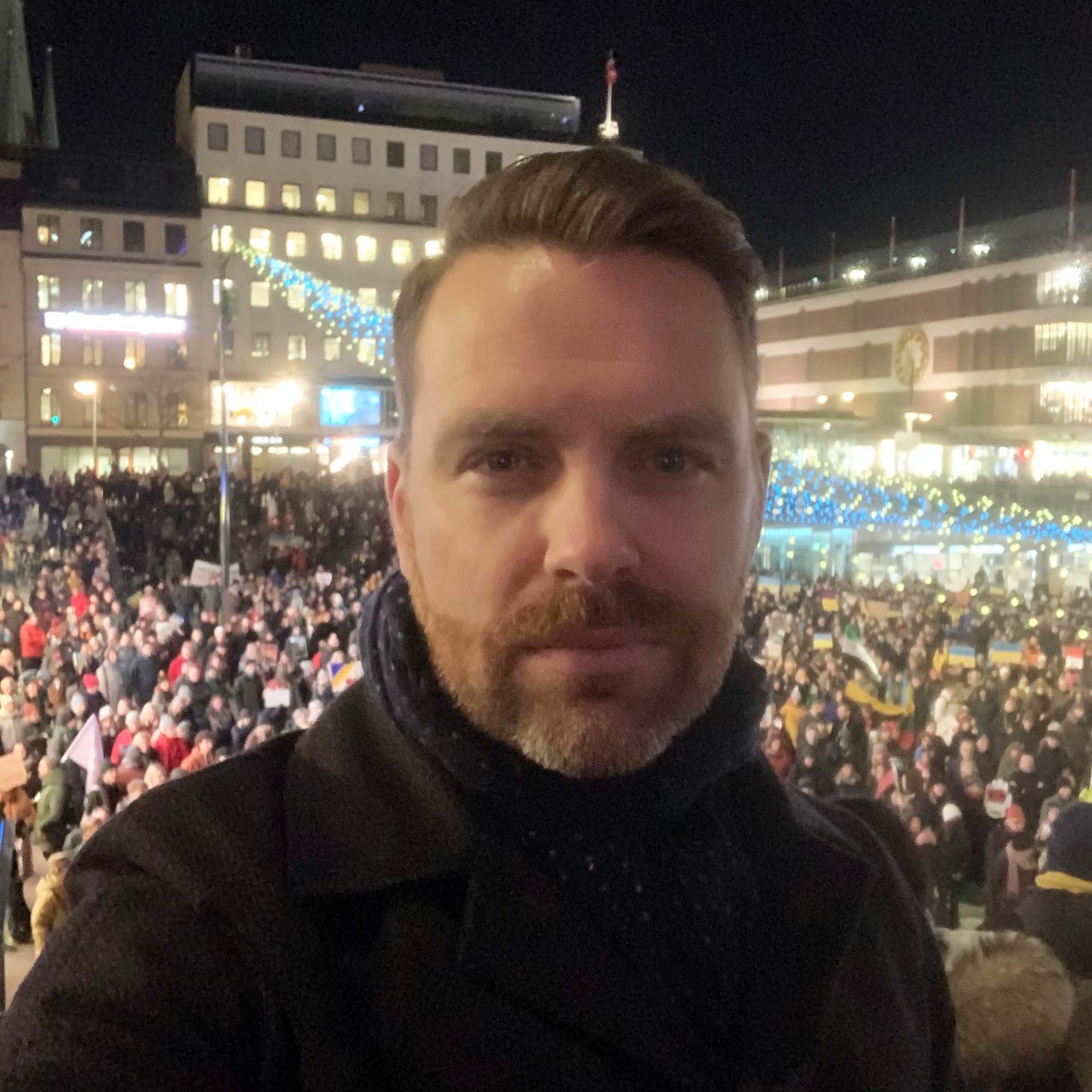 En selfie på en man med skägg i förgrunden, tagen utomhus på kvällen. Han är klädd i en mörk rock och halsduk. I bakgrunden syns en stor folkmassa på ett upplyst torg med stadshus och ljusslingor.