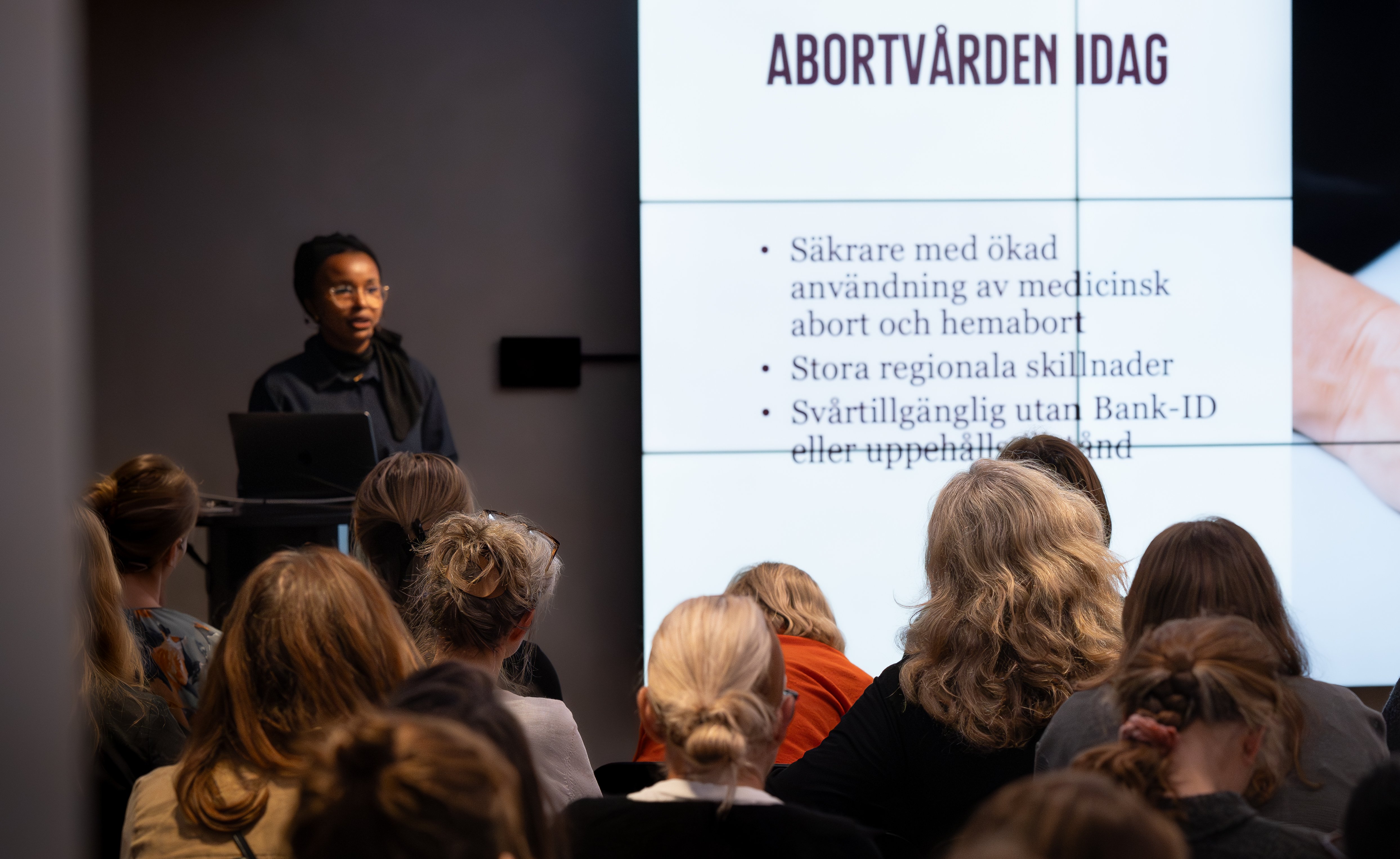 Kvinna står framför en publik med en sor skärm i bakgrunden med bokstäverna "Abortvården idag"