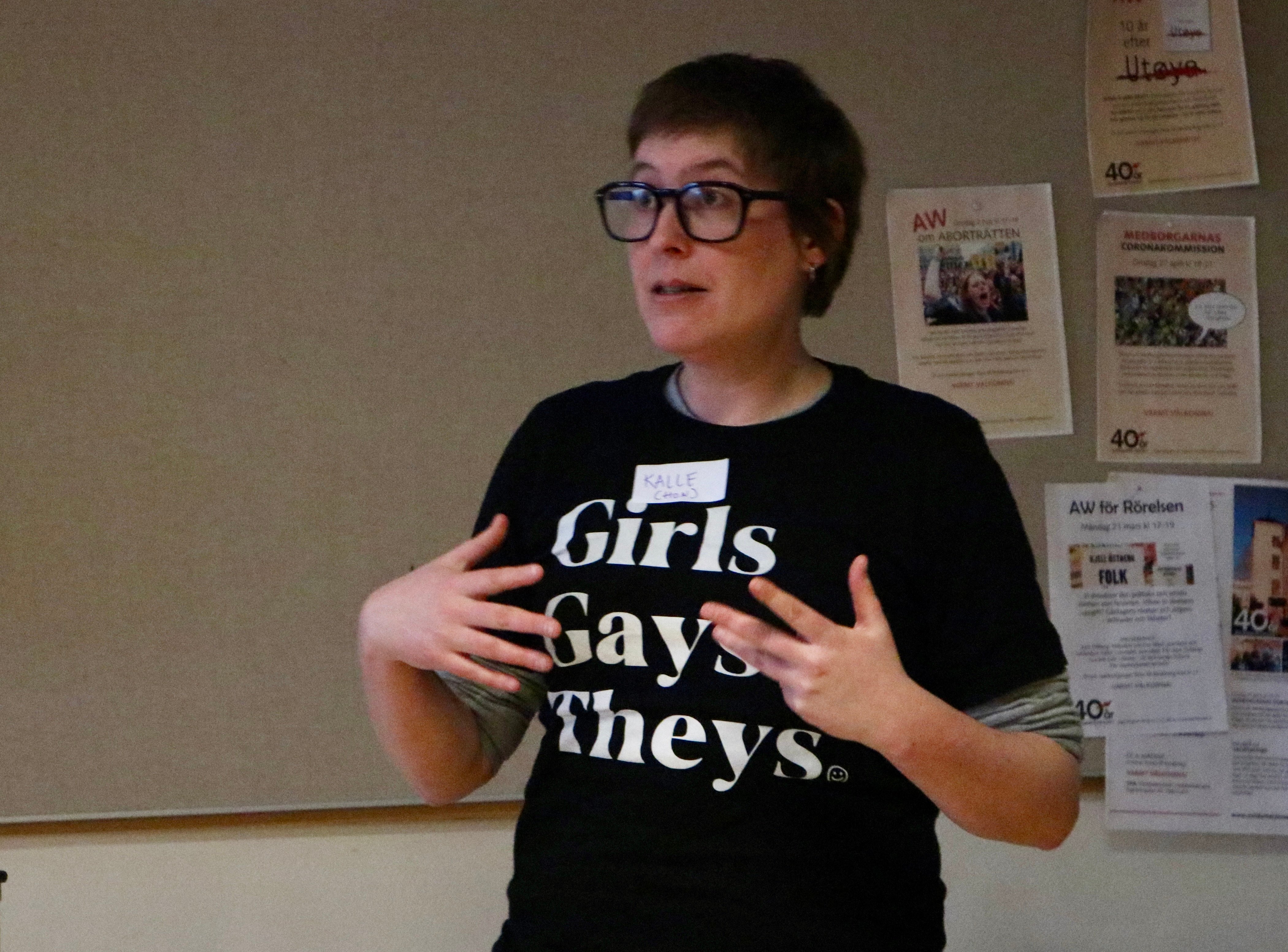 Person i glasögon och svart t-shirt med texten "Girls Gays Theys" talar och gestikulerar med händerna.