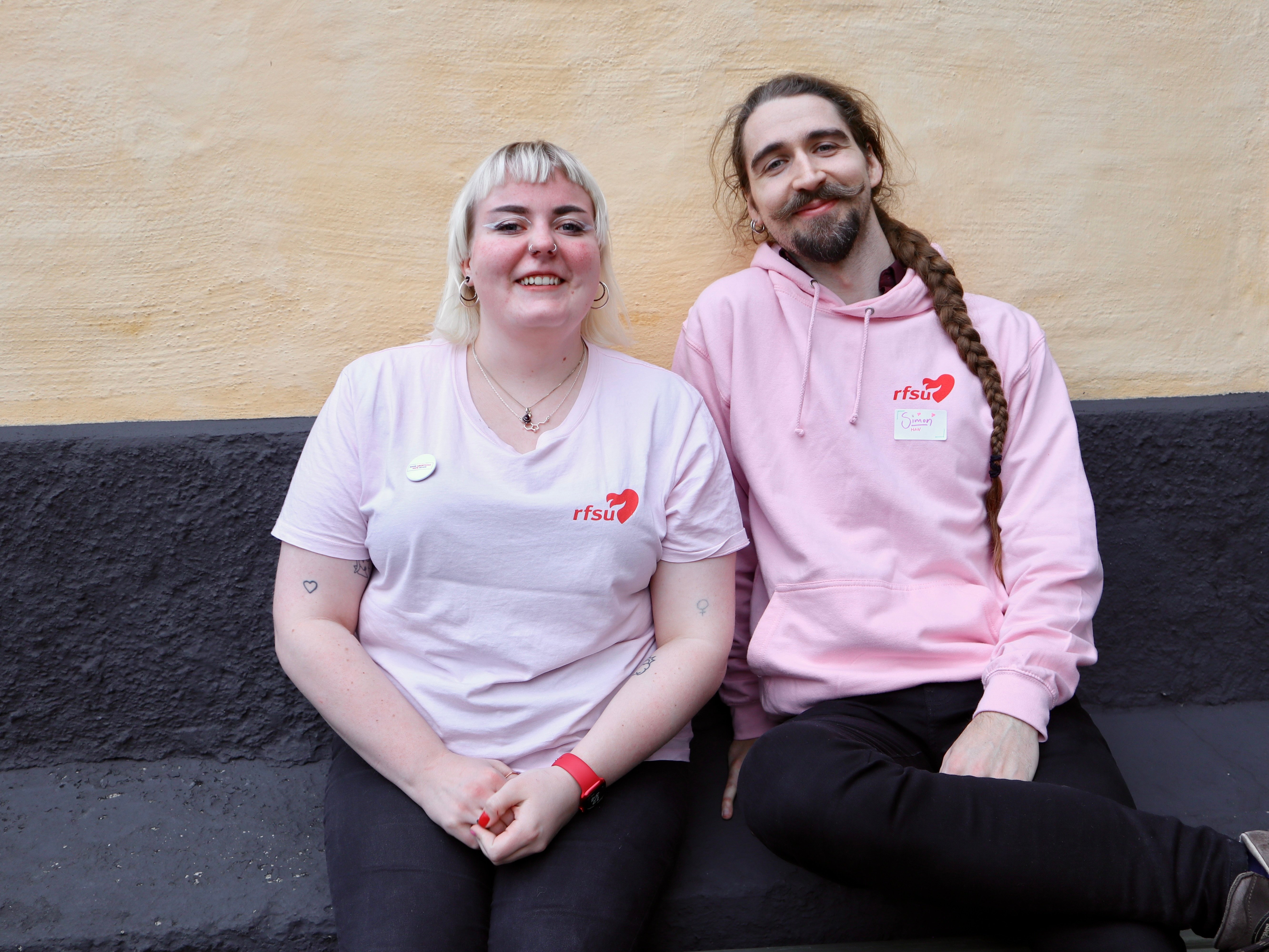 Två leende personer i rosa tröjor med RFSU-logotyp sitter tillsammans.