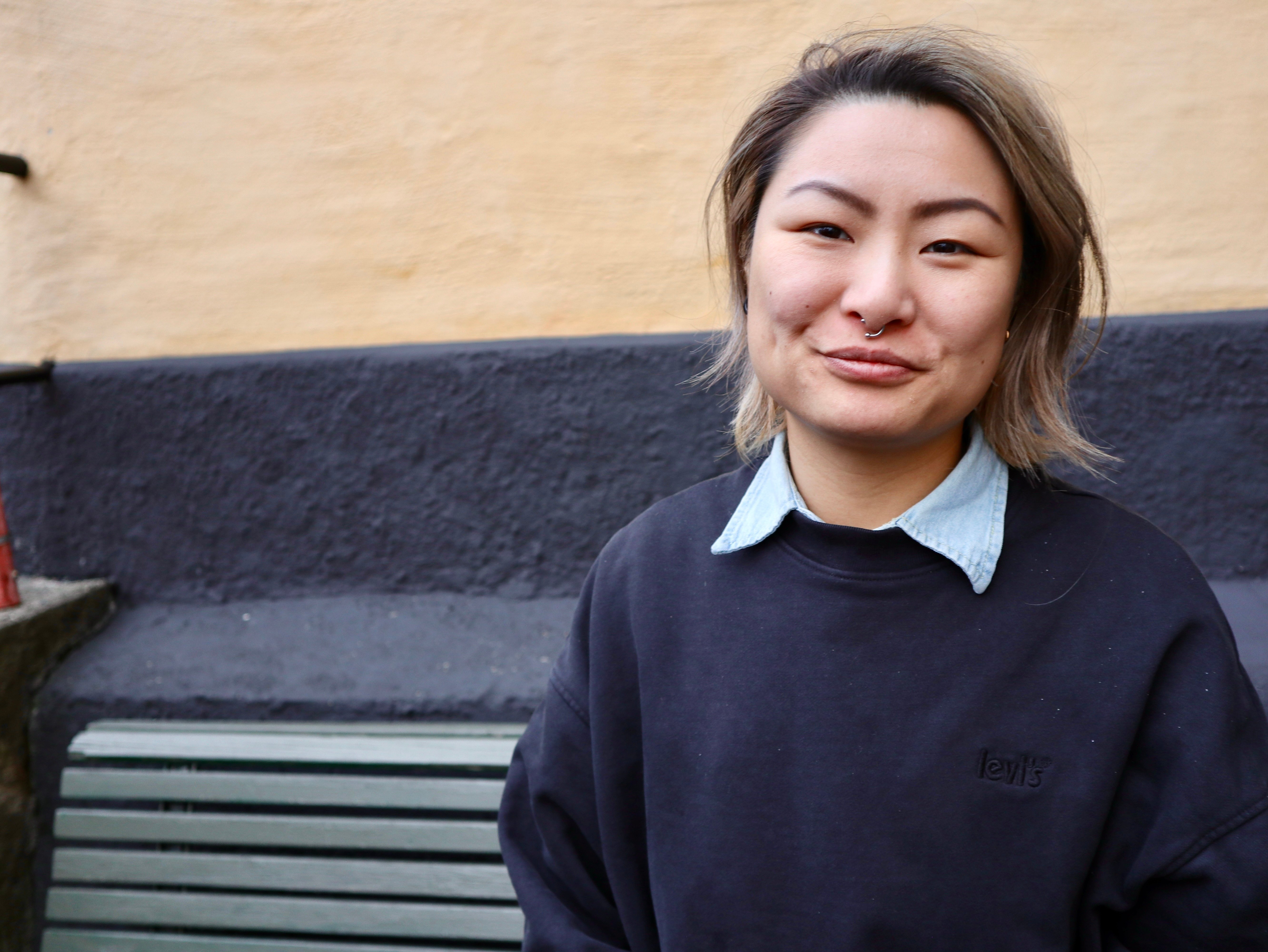 Leende person i mörkblå tröja med ljusblå krage framför en gul och mörkgrå vägg.