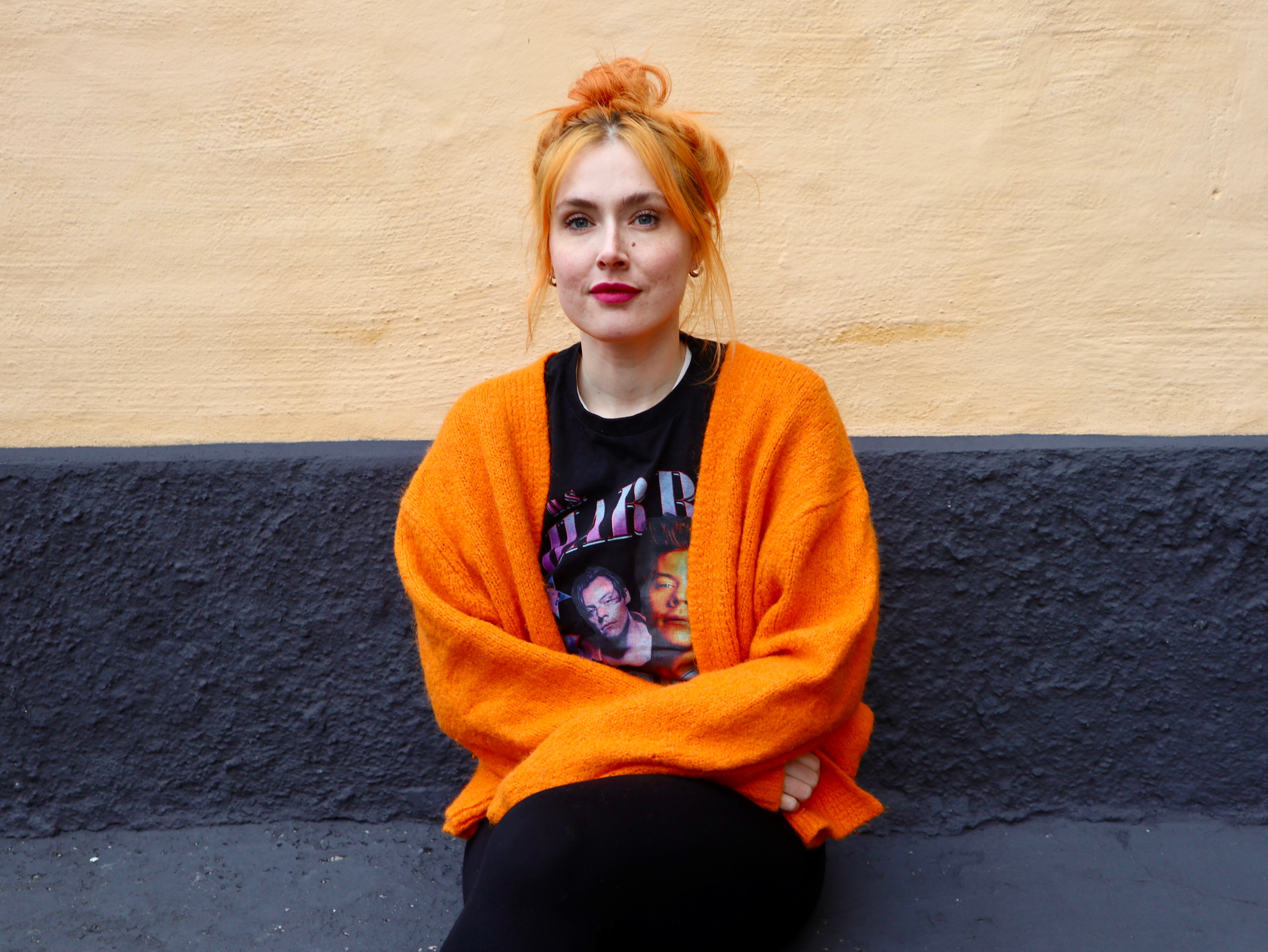 Person i orange kofta och svart t-shirt sitter mot en gul och mörkgrå vägg.
