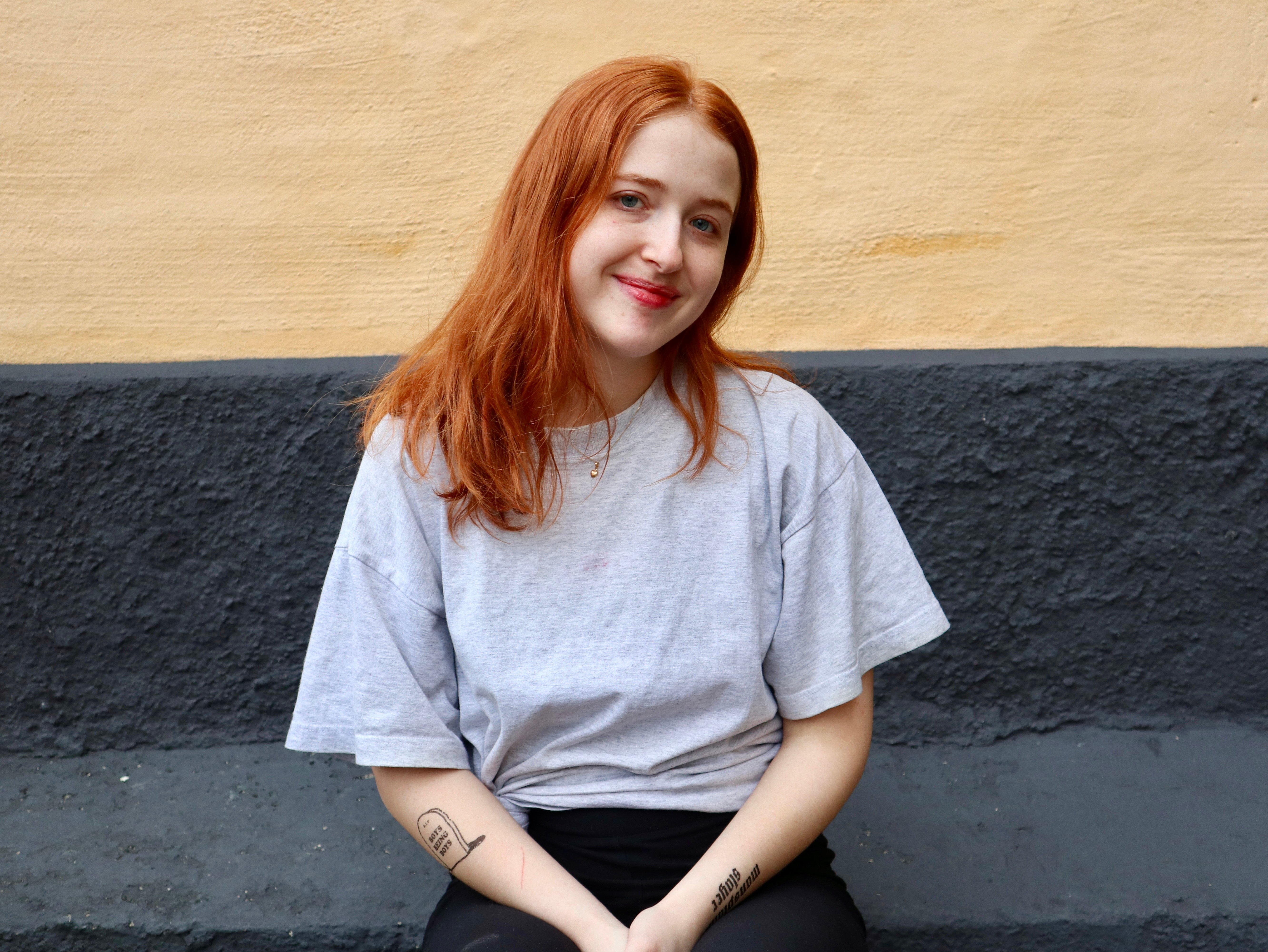 Leende person med långt rött hår och grå t-shirt sitter mot en gul och mörkgrå vägg.