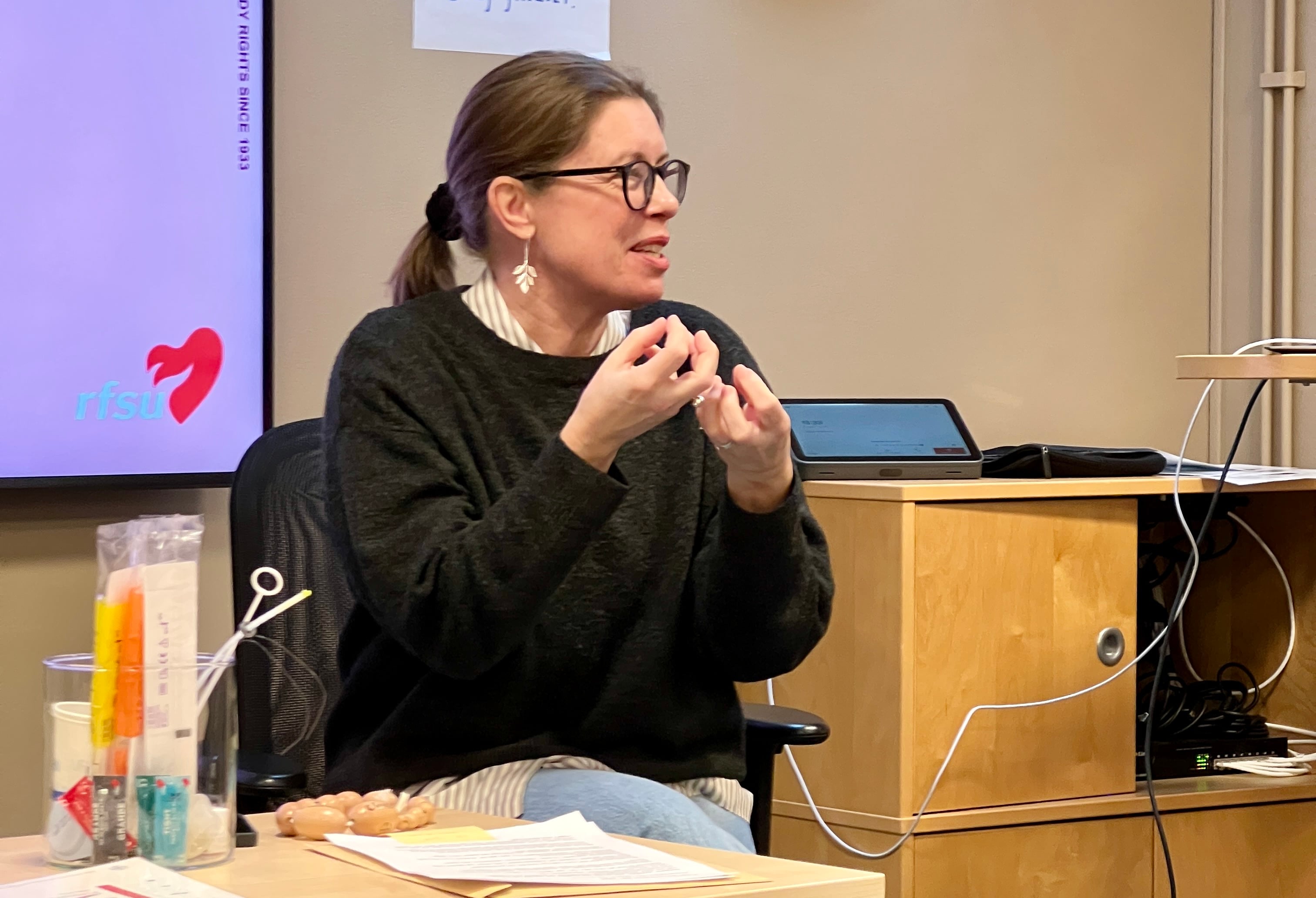 En kvinna med glasögon gestikulerar engagerat under en presentation eller workshop.
