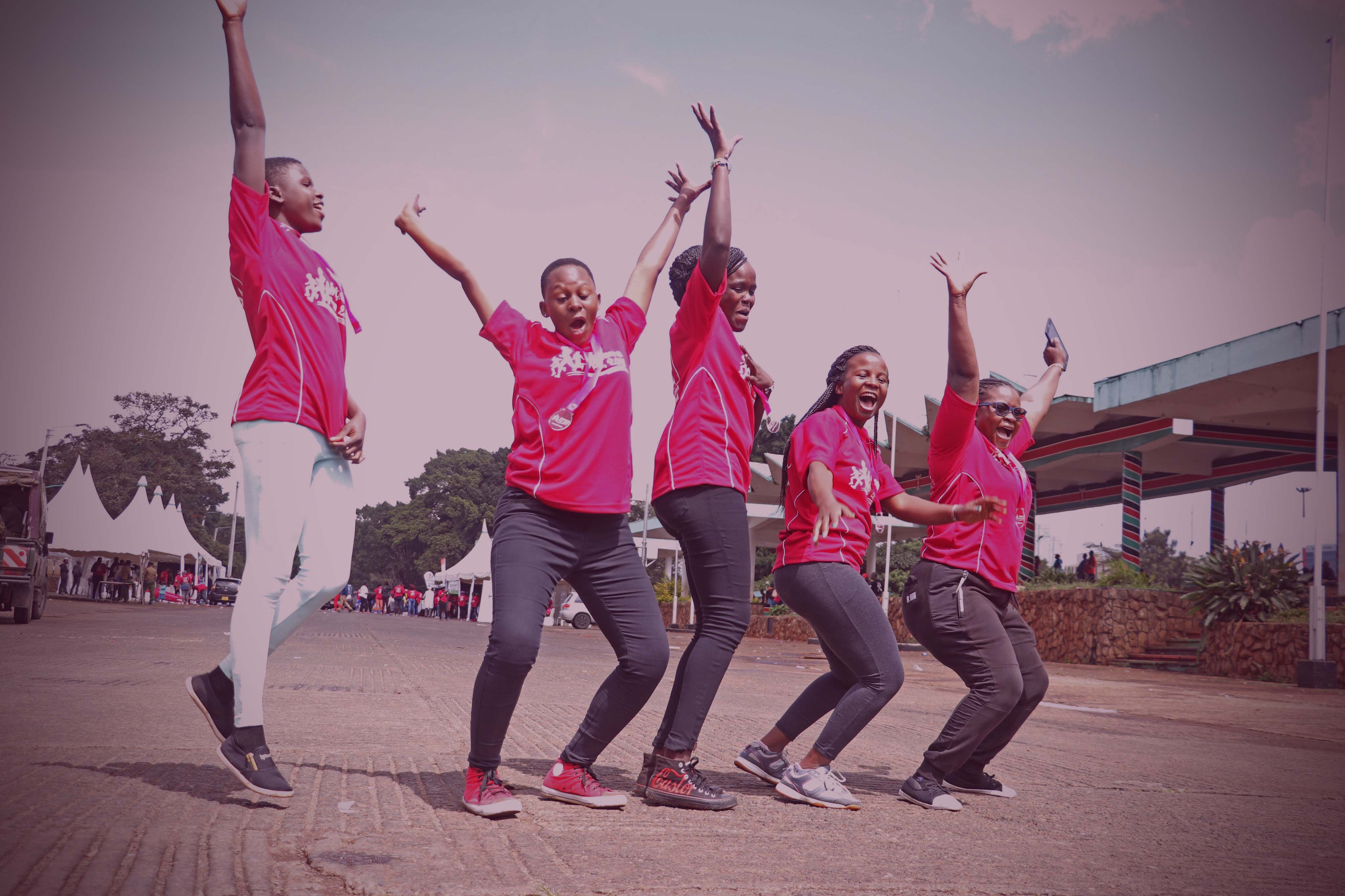 Five individuals wearing matching red t-shirts celebrate joyfully in an outdoor paved area with their arms raised in the air. One person jumps high mid-air while the others cheer, with white tents and structures visible in the background.