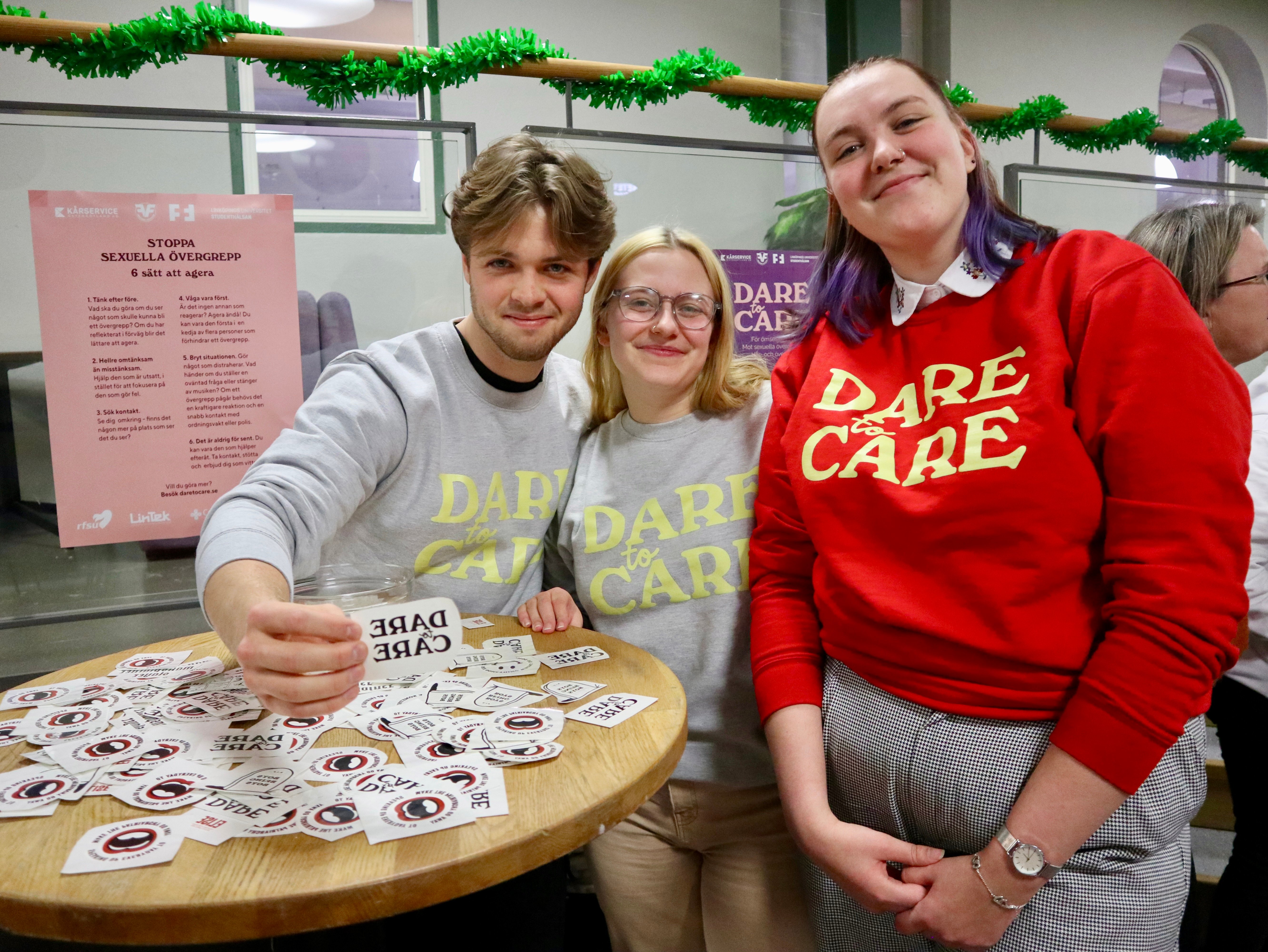 Tre ungdomar i profiltröjor står vid ett bord fyllt med klistermärken för kampanjen Dare to Care.