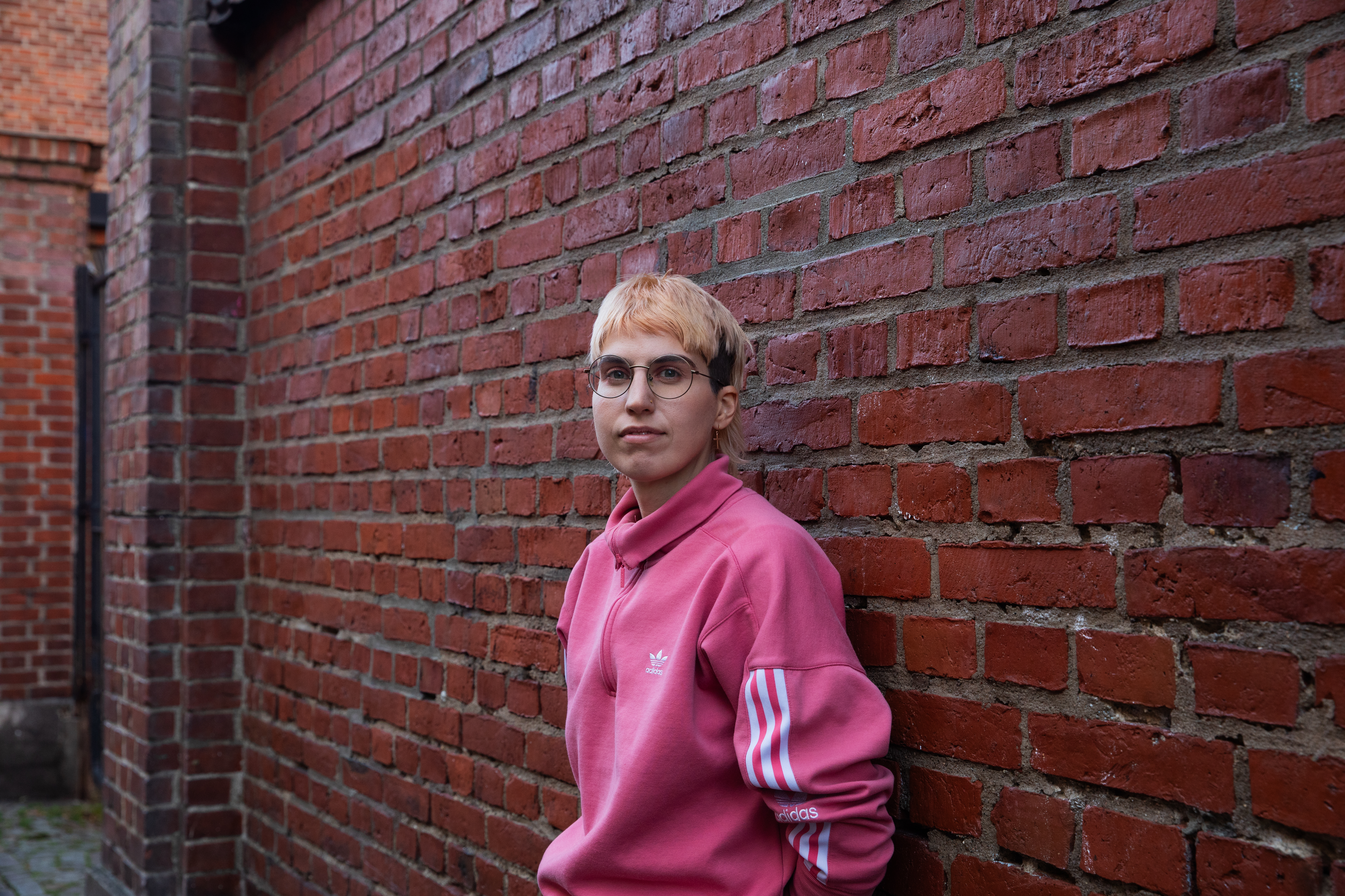 Person i rosa tröja och glasögon lutar sig mot en röd tegelvägg utomhus.