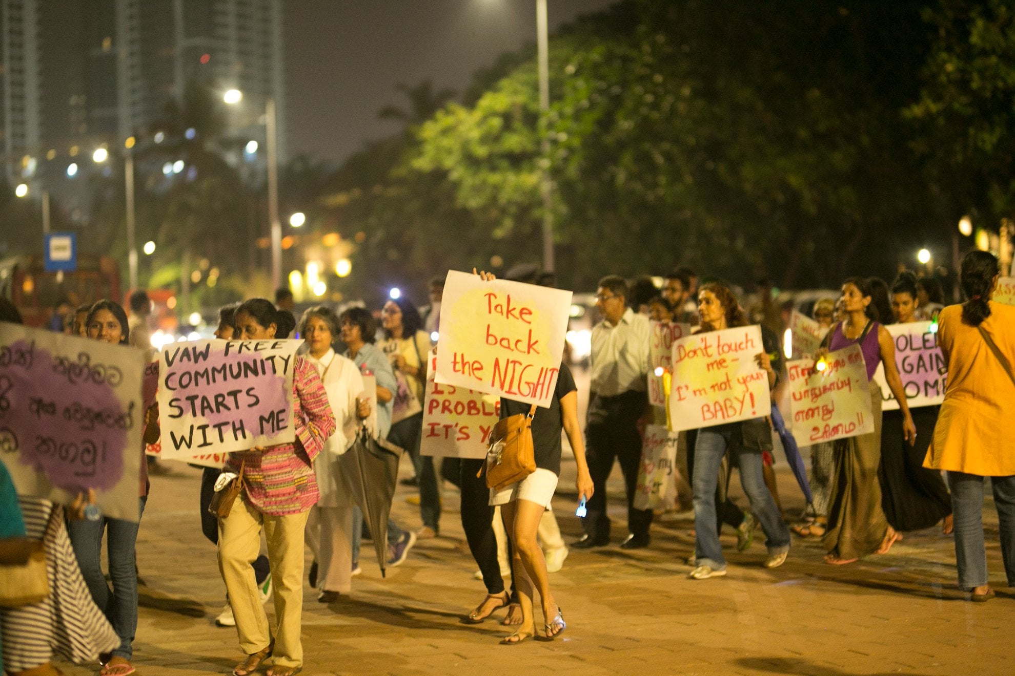 En grupp människor tågar genom en stad på natten i en "Take Back the Night"-manifestation. Deltagarna bär på handskrivna plakat med budskap som "VAW FREE COMMUNITY STARTS WITH ME", "Take back the NIGHT" och "Don't touch me – I'm not your BABY!". I bakgrunden syns stadsljus och höga byggnader under en mörk natthimmel.