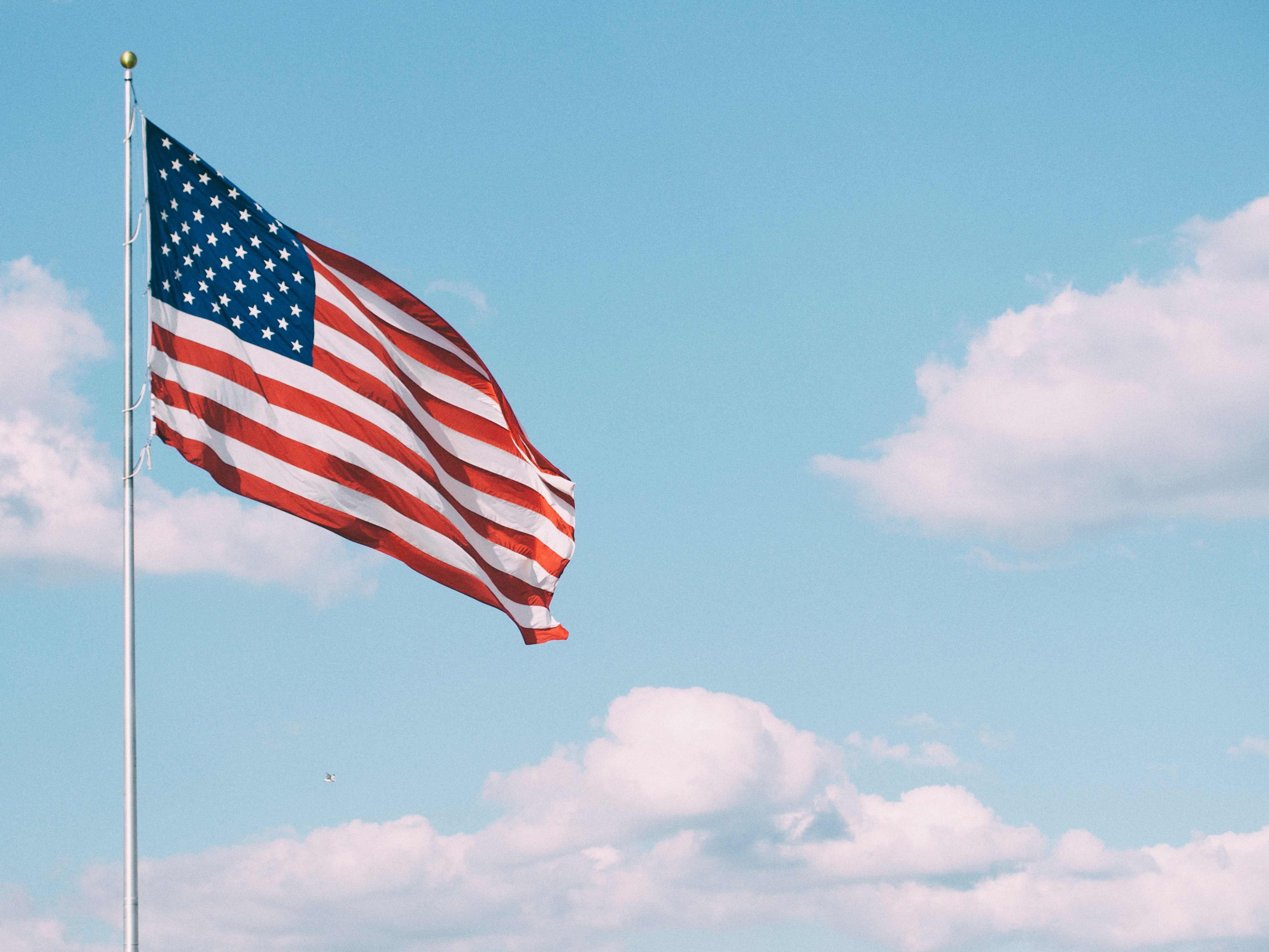The flag of the United States of America waves from a tall flagpole against a bright blue sky with scattered white clouds.