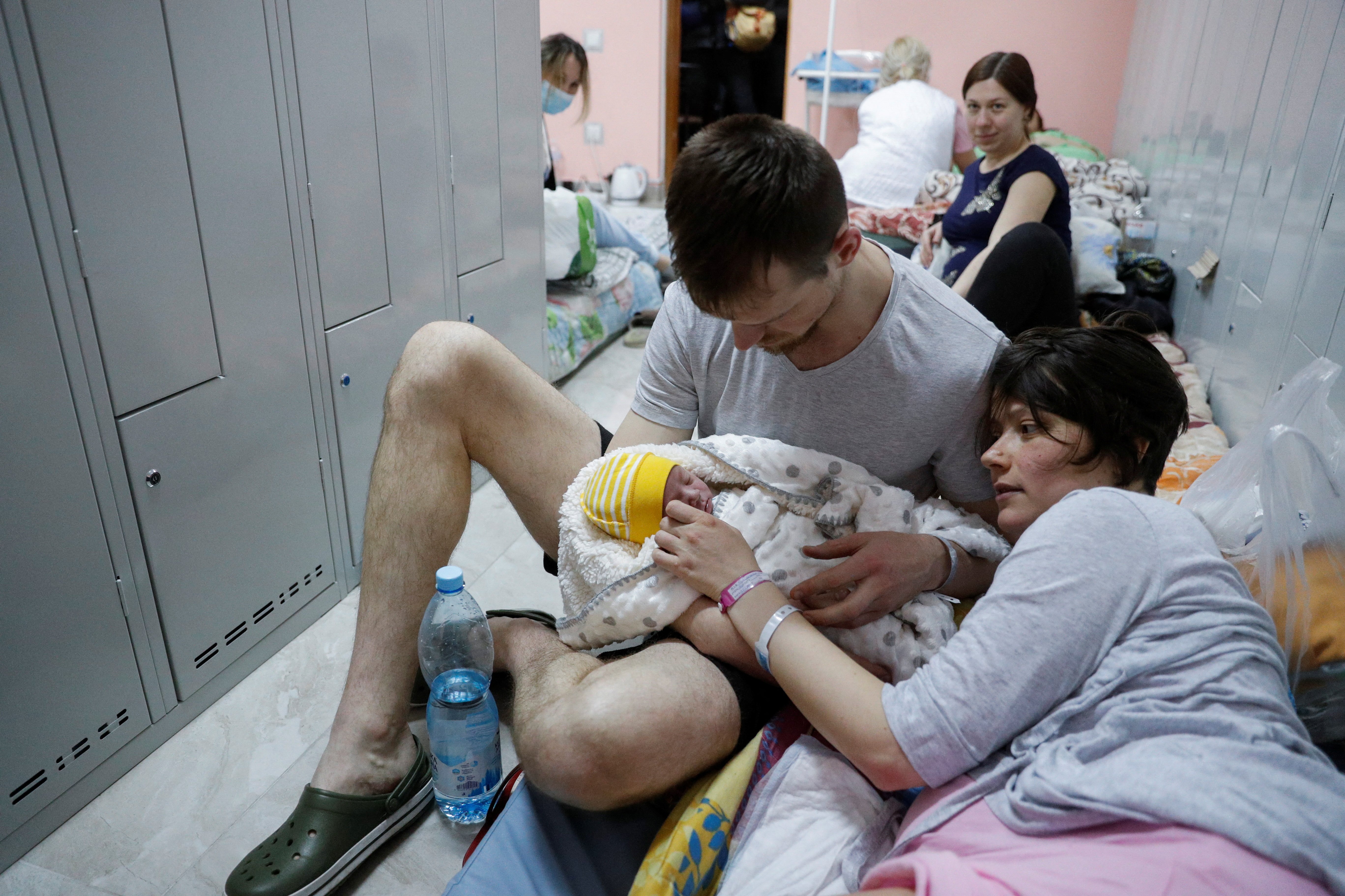 A man and a woman sit on the floor next to a row of lockers, cradling a newborn baby wrapped in a blanket. In the background, other people are seen resting on makeshift beds in a crowded, temporary shelter.