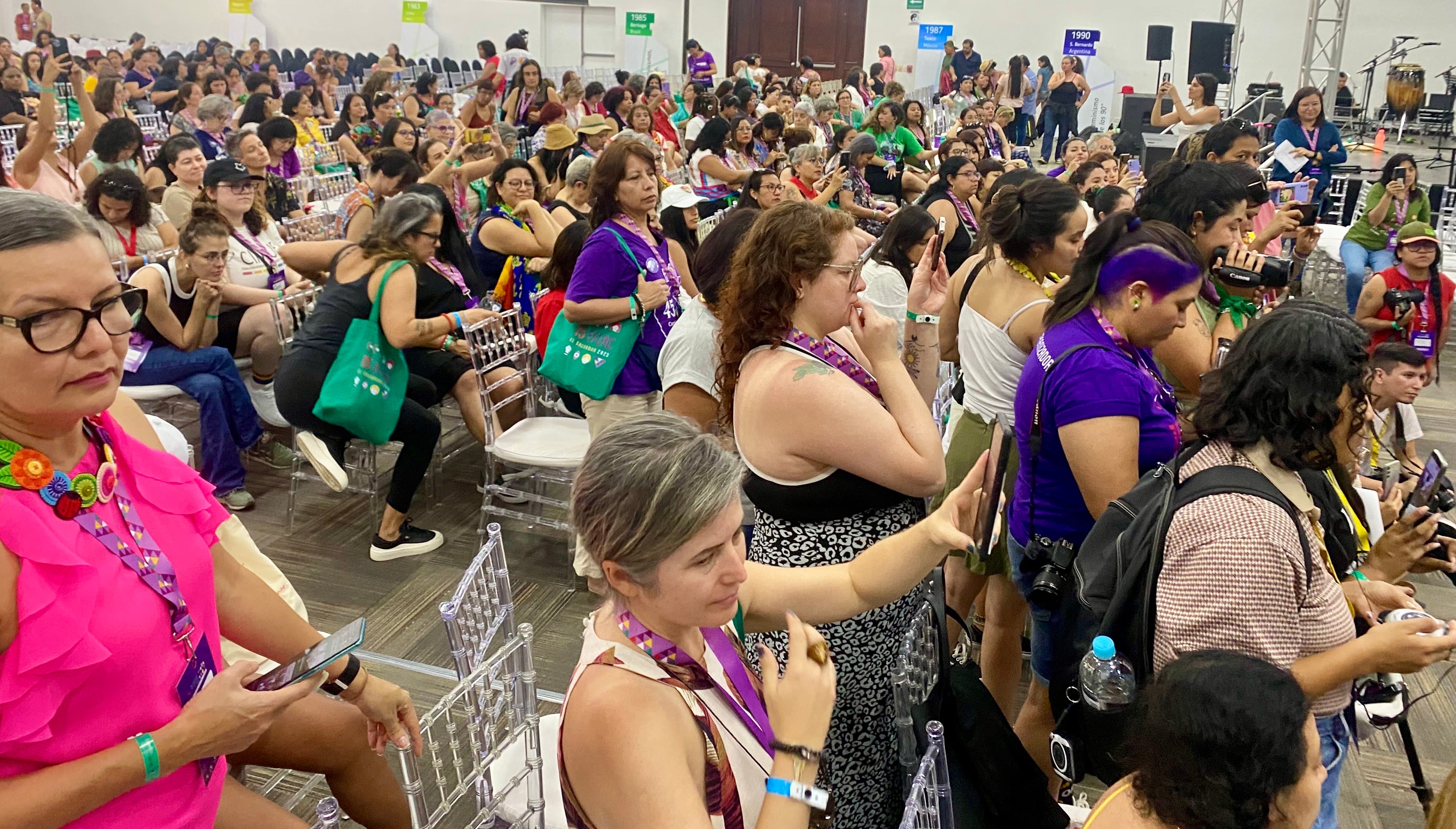 A large, crowded indoor hall is filled with people, many of whom are holding up smartphones to record or take photos. The audience members are standing or sitting in transparent chairs, with several wearing bright colors and event lanyards.