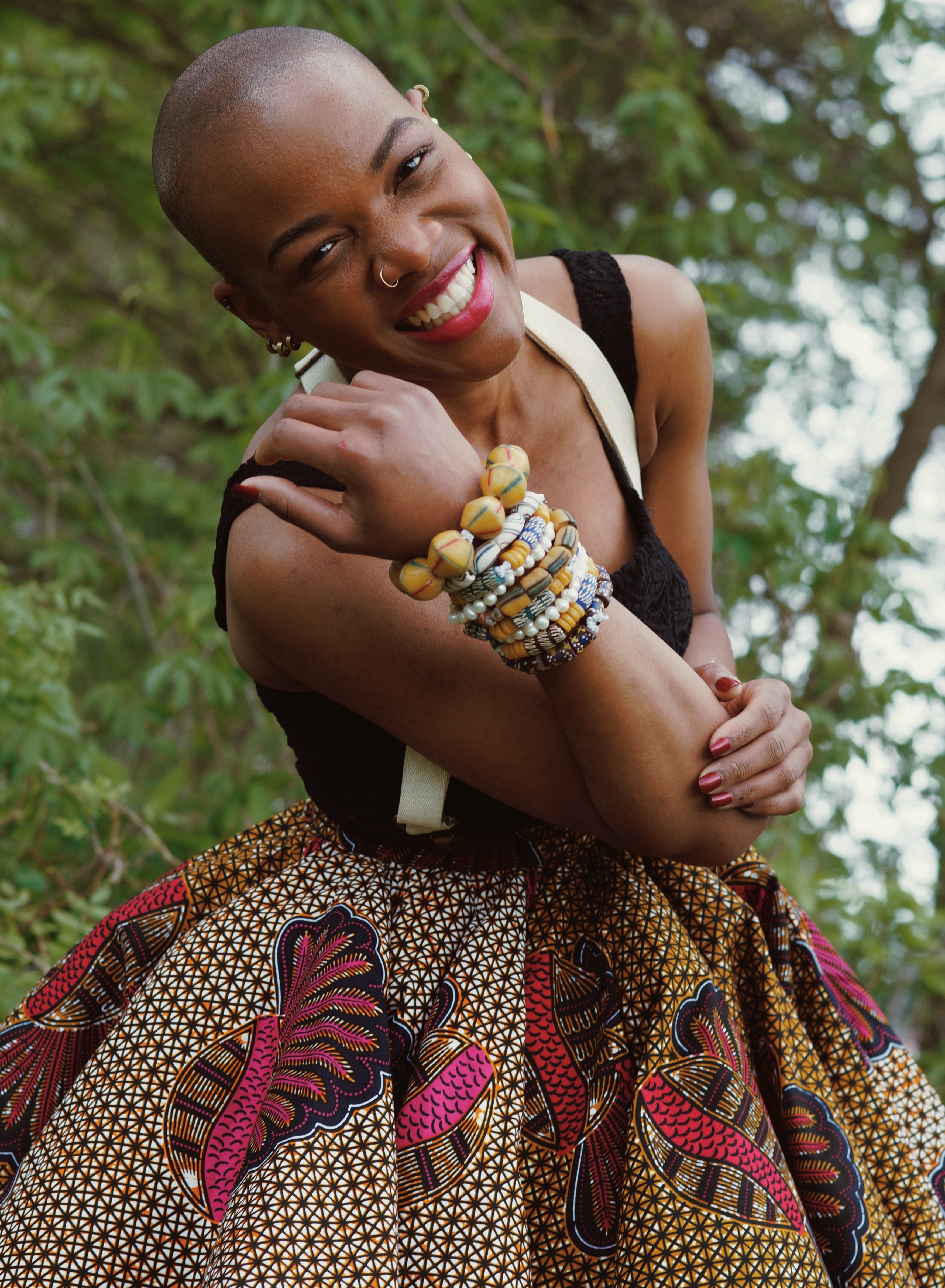 A smiling person with a shaved head leans forward in front of green foliage, wearing a vibrant, patterned full skirt and a black top. They are wearing multiple beaded bracelets on their arm, a nose ring, and bright red lipstick.