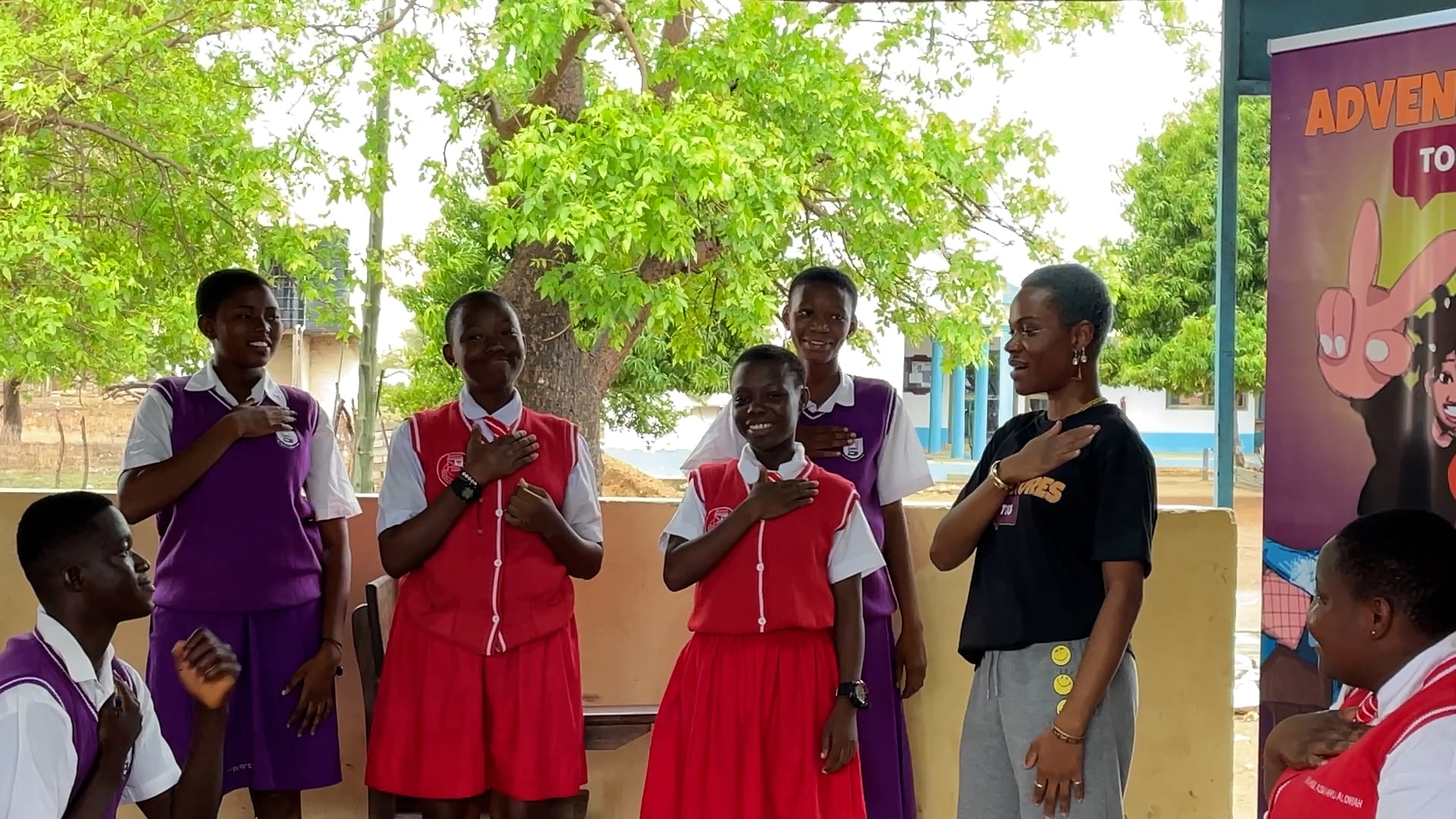 A group of individuals stand in a line under a covered outdoor area, with several wearing red or purple uniforms and placing their right hands over their hearts. A woman in a black t-shirt stands alongside them, mirroring the gesture.