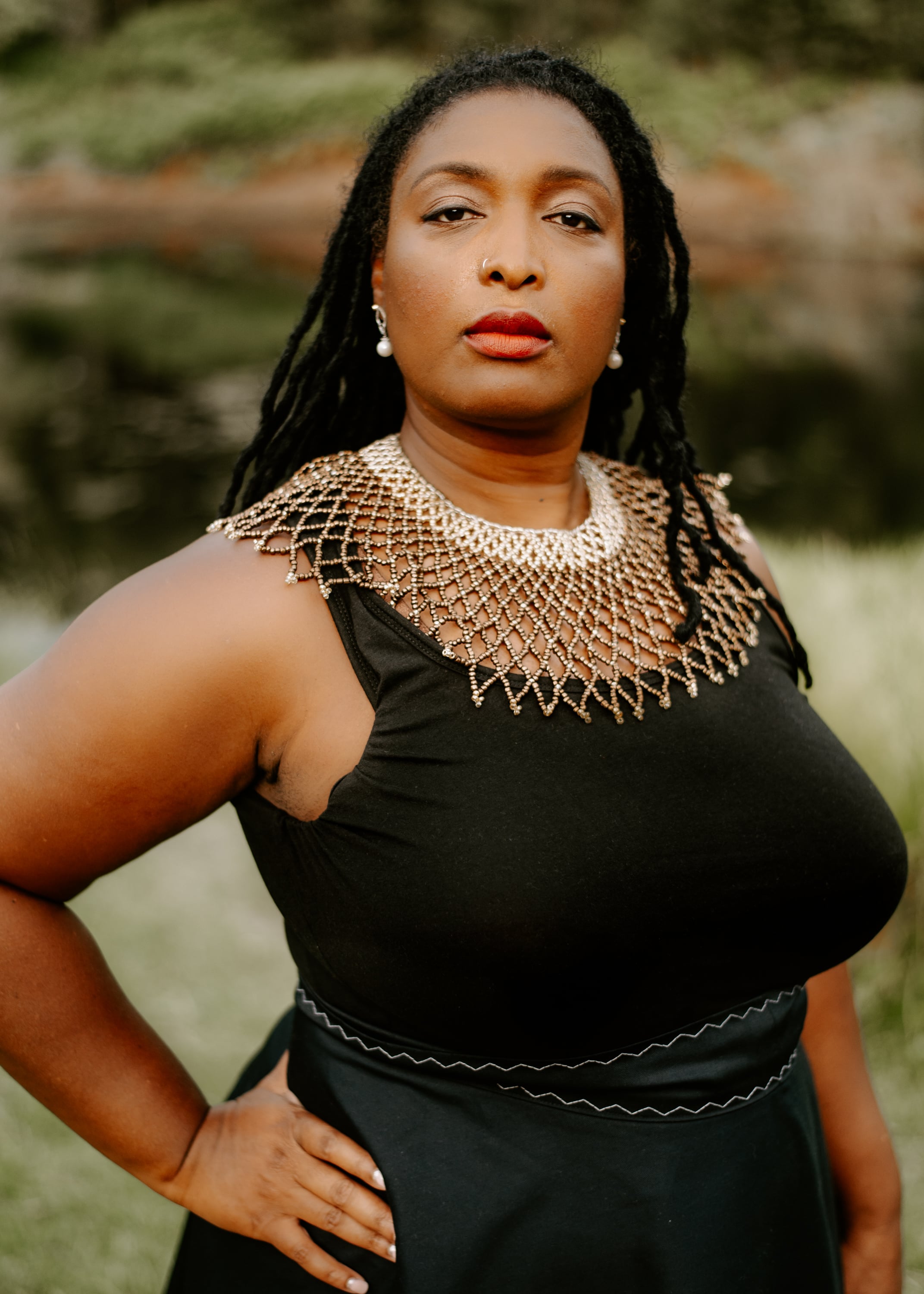 A woman with long locs looks directly at the camera with a poised expression, posing with one hand on her hip outdoors. She is wearing a black top accented by an intricate, wide beaded gold collar necklace.