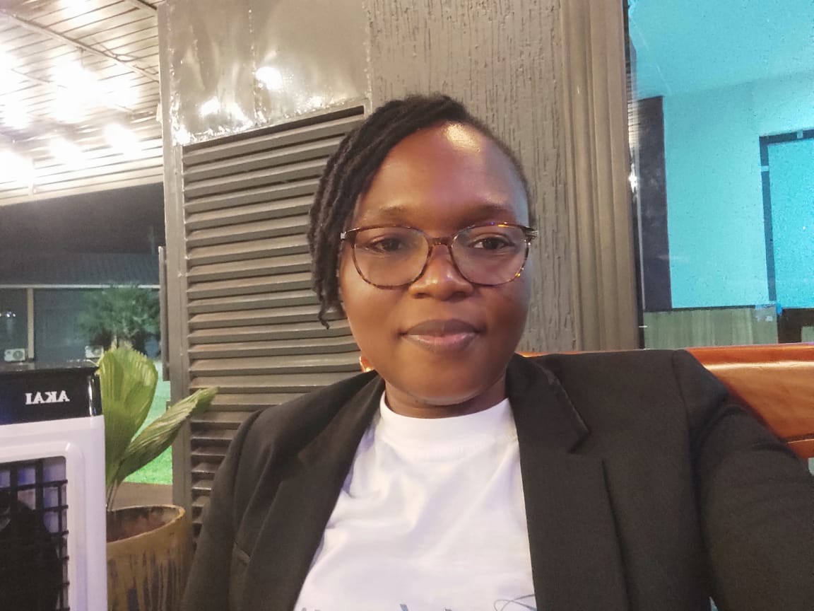 A person wearing glasses, a white t-shirt, and a black blazer looks toward the camera with a soft smile. They are seated indoors with a structured dark wall and ambient lighting in the background.
