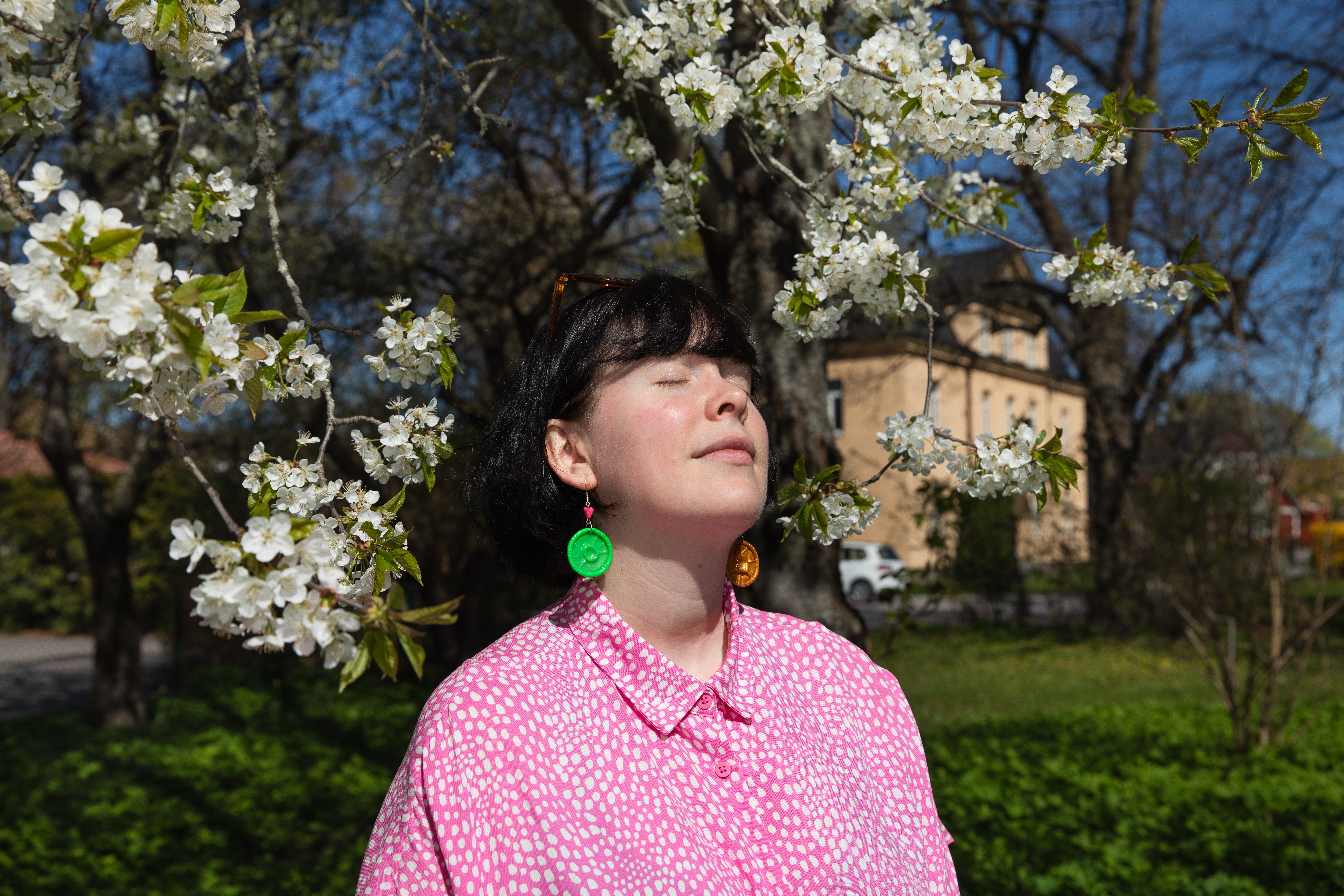 En person i rosa mönstrad blus står utomhus med slutna ögon under ett blommande körsbärsträd. Personen bär stora, runda örhängen i grönt och orange, och solljuset silar ner genom de vita blommorna.