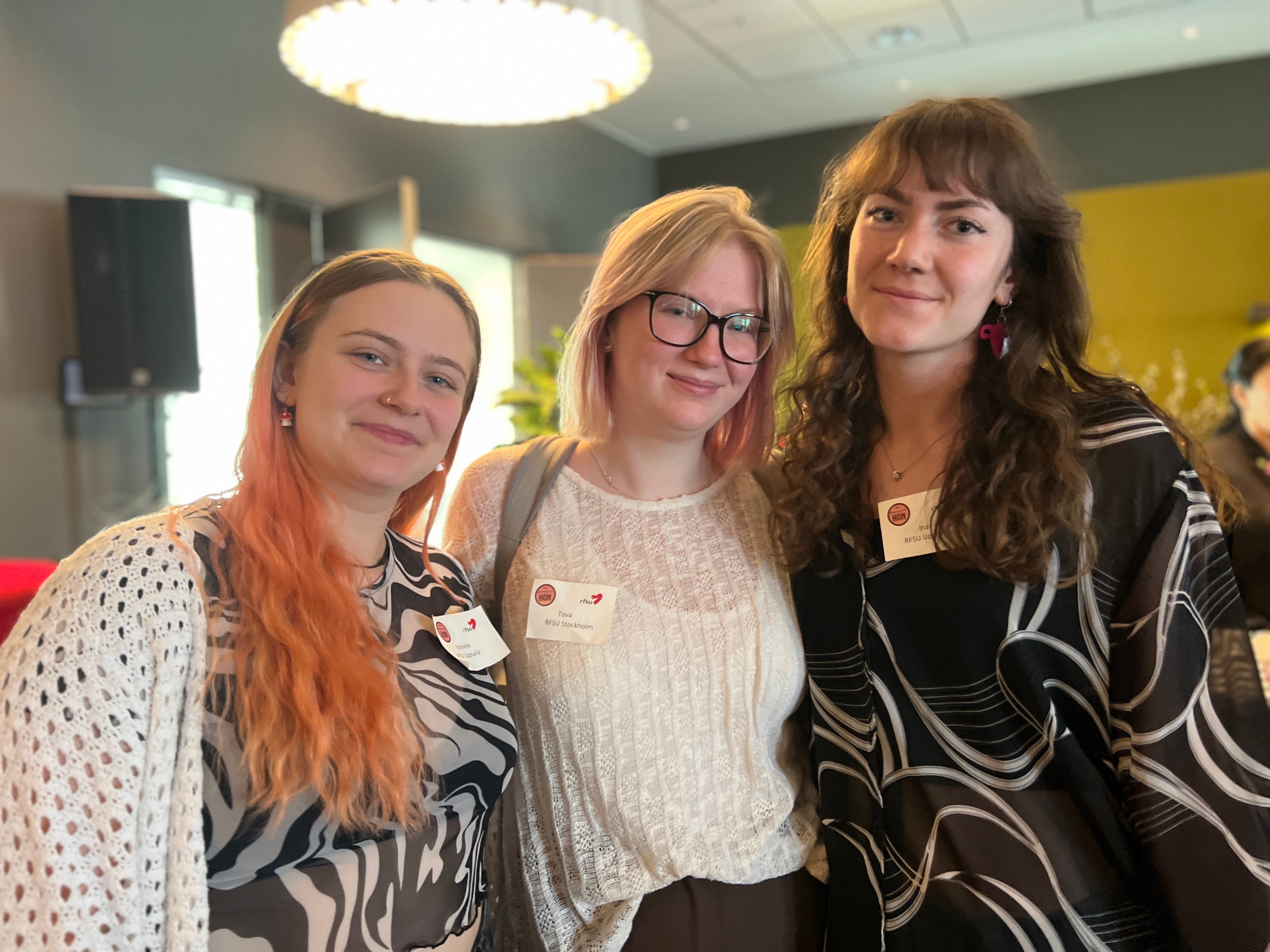 Tre unga kvinnor poserar tillsammans och ler mot kameran i en konferensmiljö.