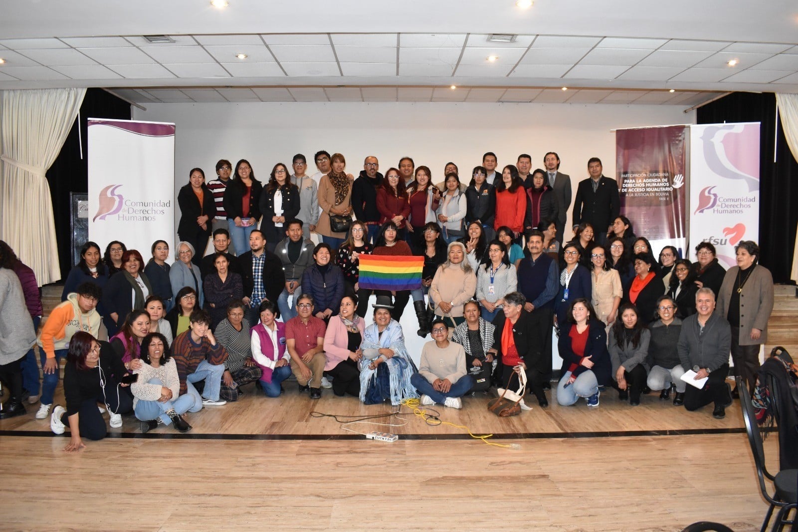 A large group of people pose together for a photograph on a stage, with an individual in the center holding up a rainbow pride flag. Large informational banners flank the group on both sides.