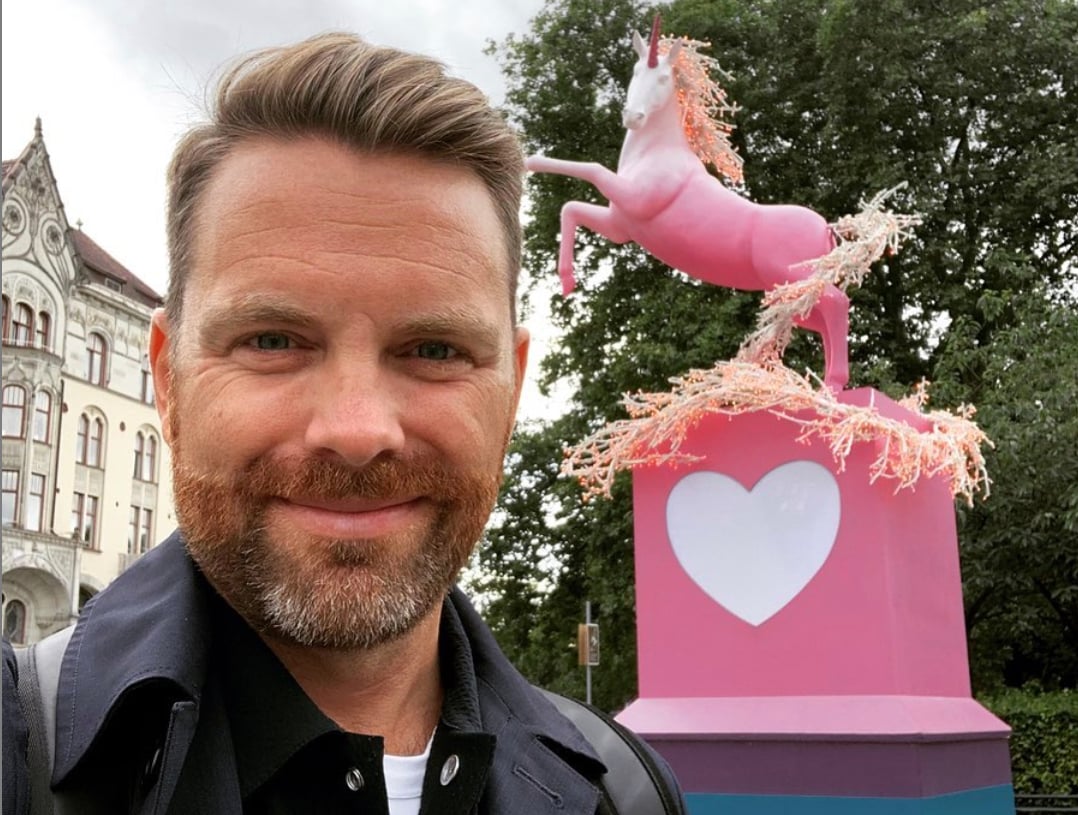 En närbild i form av en selfie på en leende man med rödbrunt skägg och välkammat hår. Han står utomhus med en stor rosa skulptur i bakgrunden som föreställer en enhörning på en piedestal dekorerad med ett vitt hjärta. I bakgrunden till vänster syns en vacker, äldre byggnad med utsmyckad fasad, och till höger skymtar gröna träd.