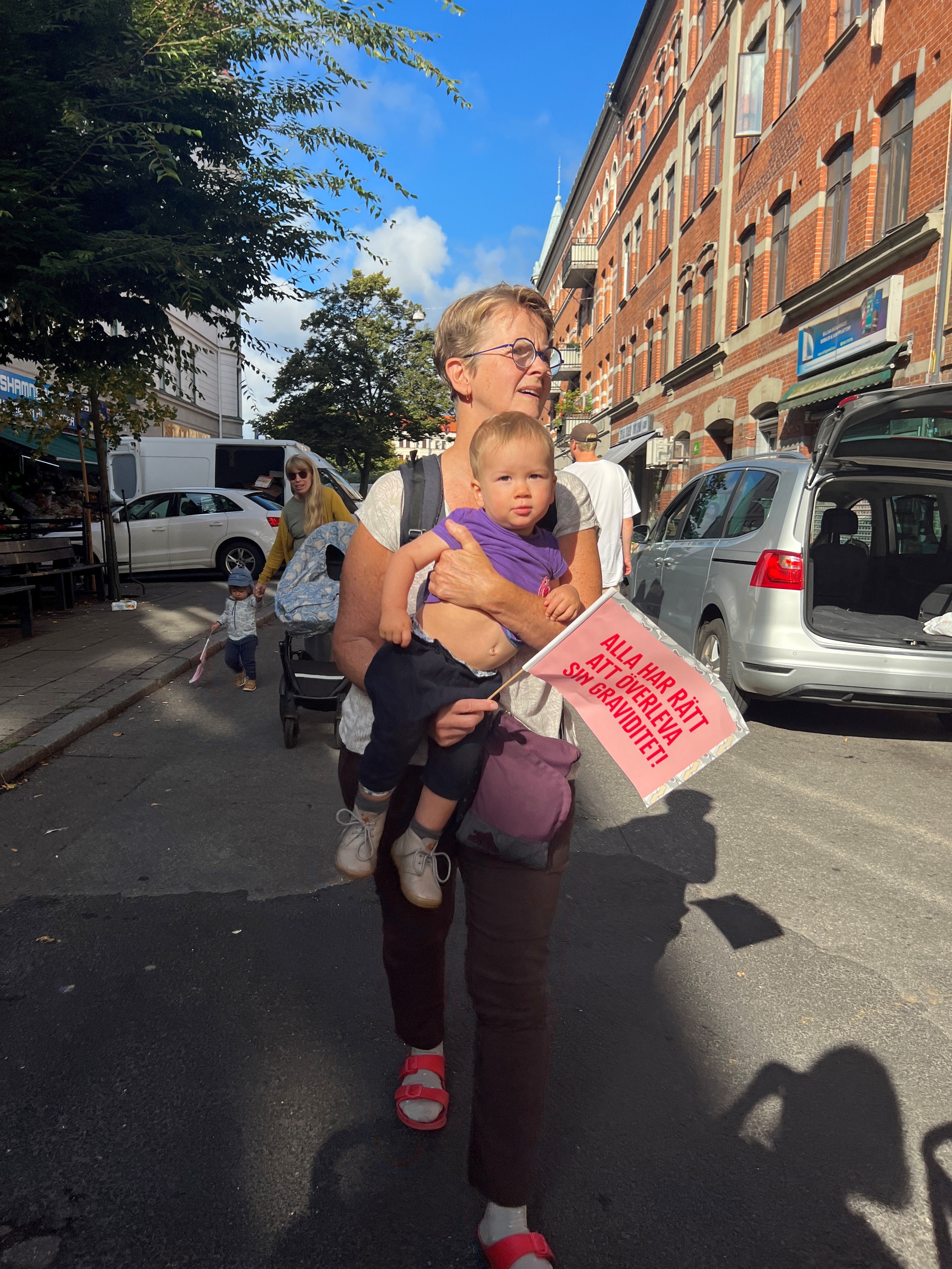 En person håller i ett barn som håller i en flagga utomhus i solen