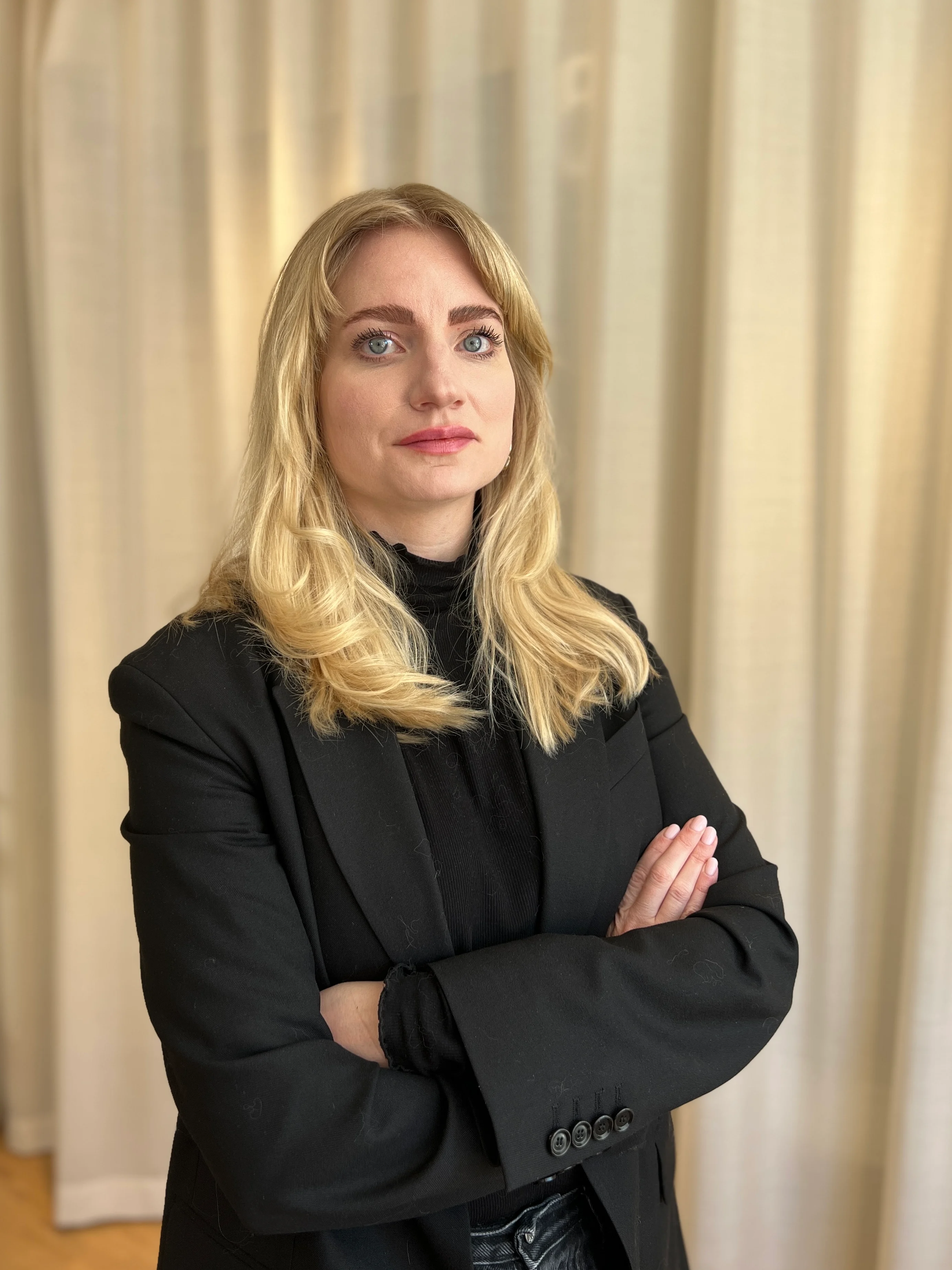 Pressbild på en kvinna med blondt hår i svart kavaj som står med armarna i kors mot en ljus gardinbakgrund.