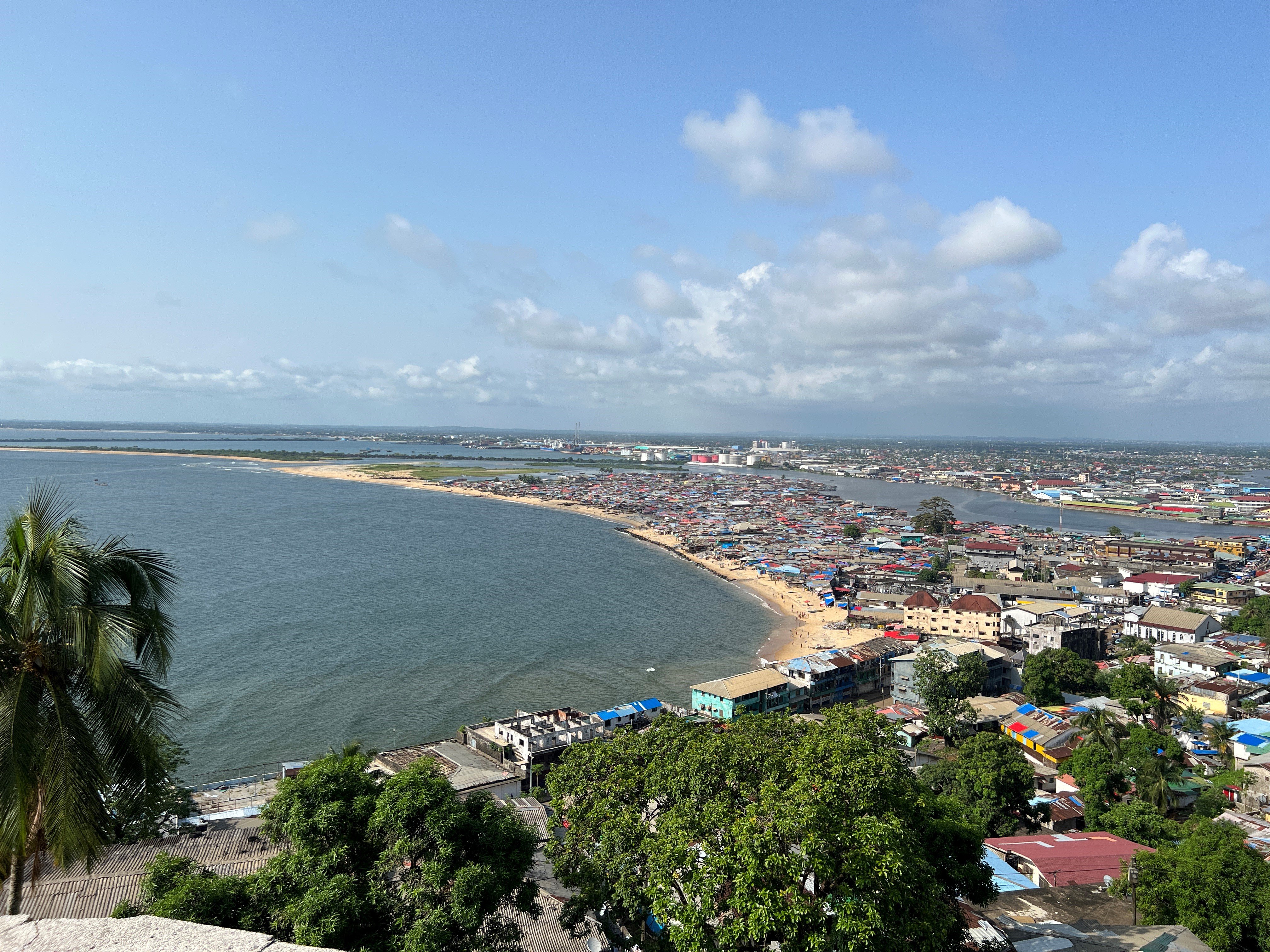 A high-angle panoramic view shows a densely populated coastal settlement situated on a curved sandy peninsula. The community is bordered by open blue water on one side and a river inlet on the other under a bright sky.