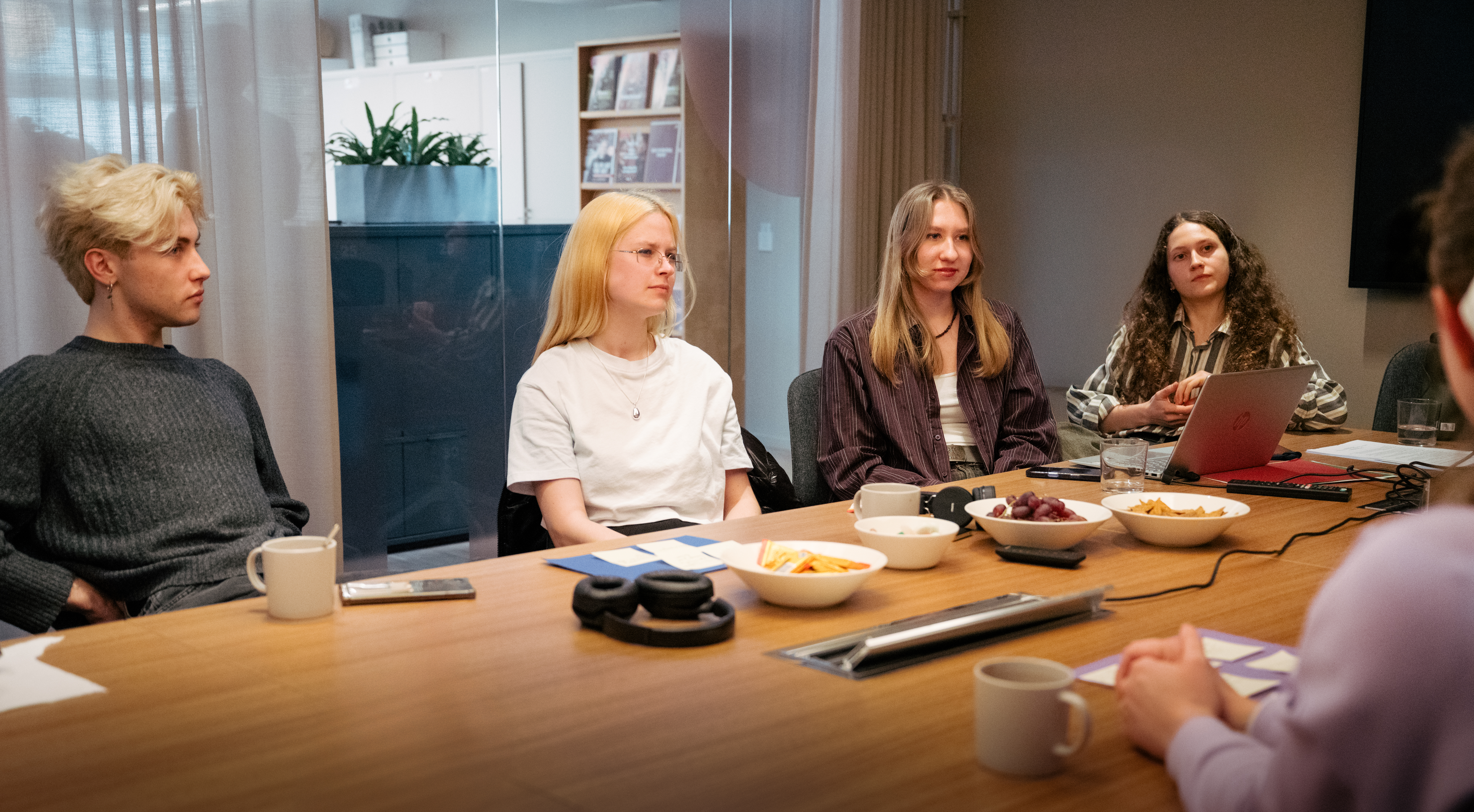 Fyra personer sitter längs ena sidan av ett långt mötesbord i ett konferensrum. På bordet står skålar med snacks, kaffekoppar och hörlurar.