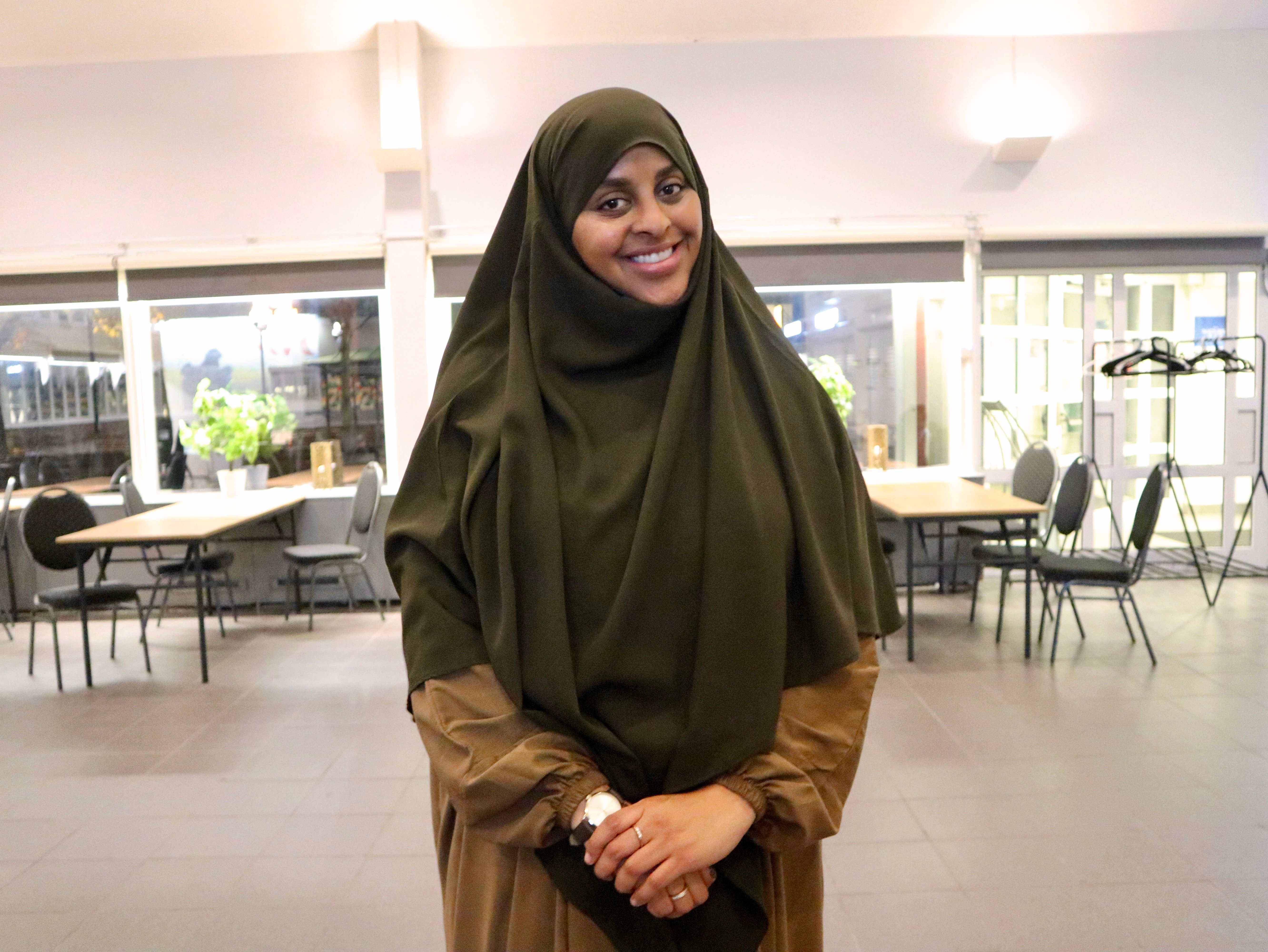 Leende person i mörkgrön slöja och brun klädnad står i en ljus lokal.