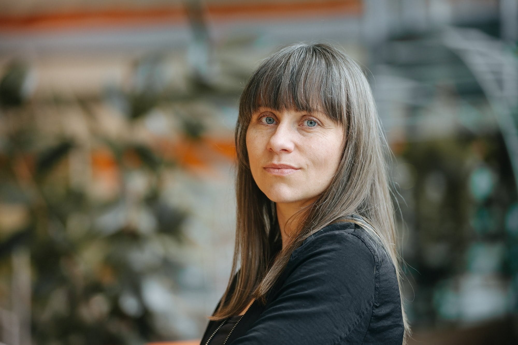 Kvinna med svart tröja långt hår och lugg står snett och tittar mot kameran