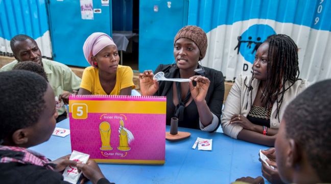 En grupp unga sitter runt ett bord och tittar när en person visarupp en kondom under ett möte om familjeplanering och sexuell och reproduktiv hälsa, foto.  