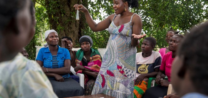 En grupp kvinnor från Pakamus kvinnogrupp i Uganda lyssnar på föreläsning om familjeplanering. En föreläsande kvinna står i mitten och håller upp en kondom.  