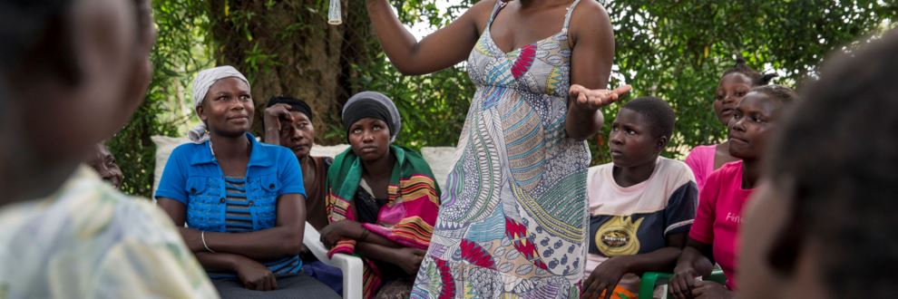 En grupp kvinnor från Pakamus kvinnogrupp i Uganda lyssnar på föreläsning om familjeplanering. En föreläsande kvinna står i mitten och håller upp en kondom.  
