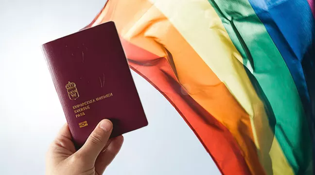 En hand som håller i ett svenskt pass och en regnbågsflagga i bakgrunden, fotografi/collage. 