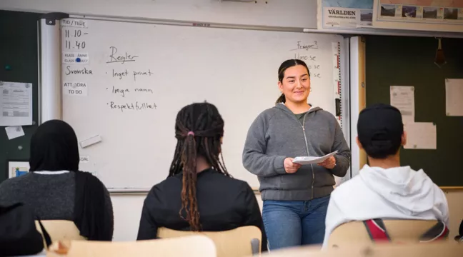 En kvinna står vid tavlan i ett klassrum framför tre elever. 