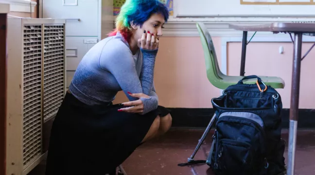 En person med regnbågsfärgat hår och långa naglar sitter på huk och ser fundersam ut i en lektionssal 
