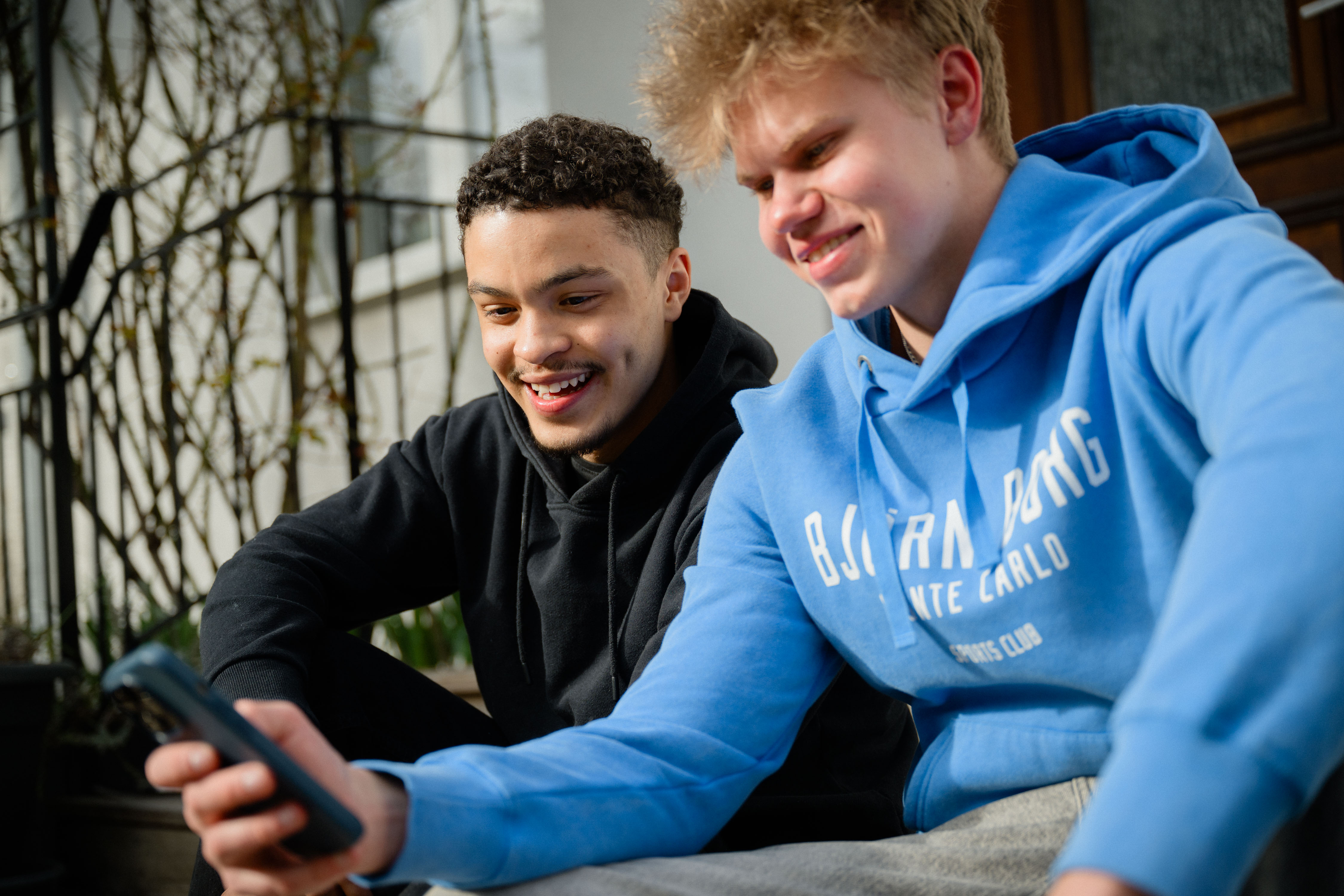 Två unga män sitter på en trappa och tittar på en telefon och ler 