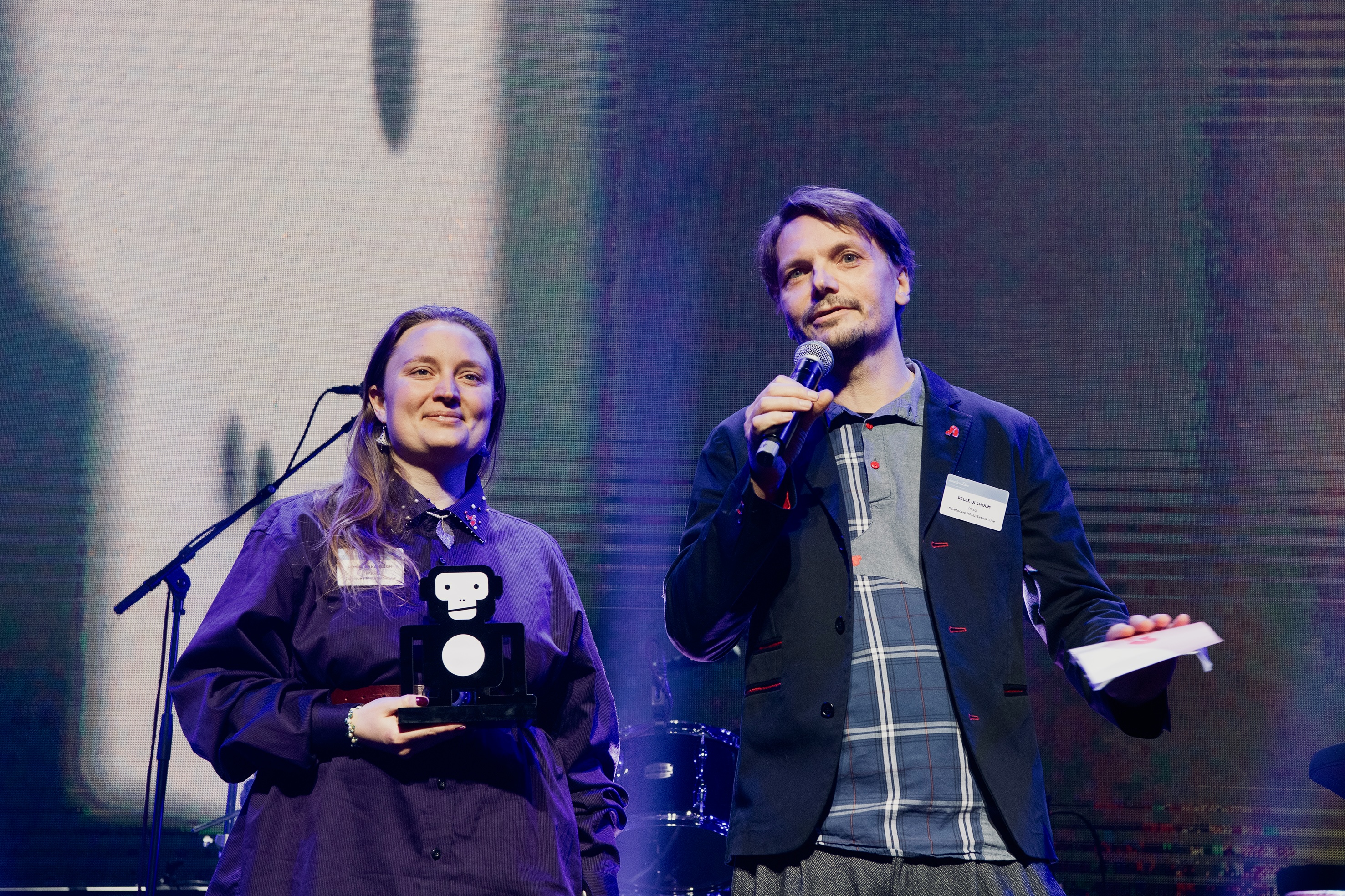 Två personer står på en scen med ett pris och pratar i en mikrofon 