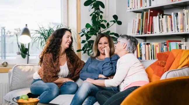 Tre kvinnor i medelåldern sitter nära varandra och skrattar i en soffa 