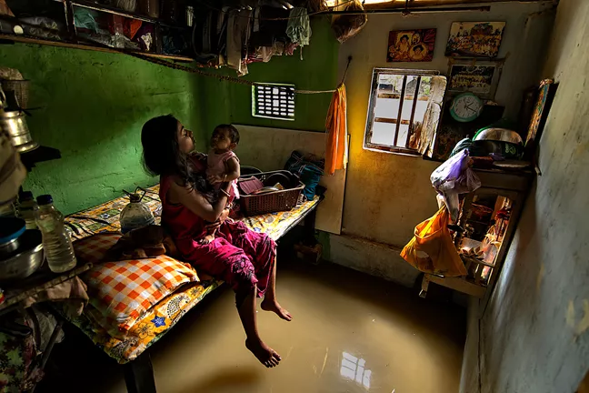 Woman sitting on the edge of a bed holding a baby. The floor is flooded.  