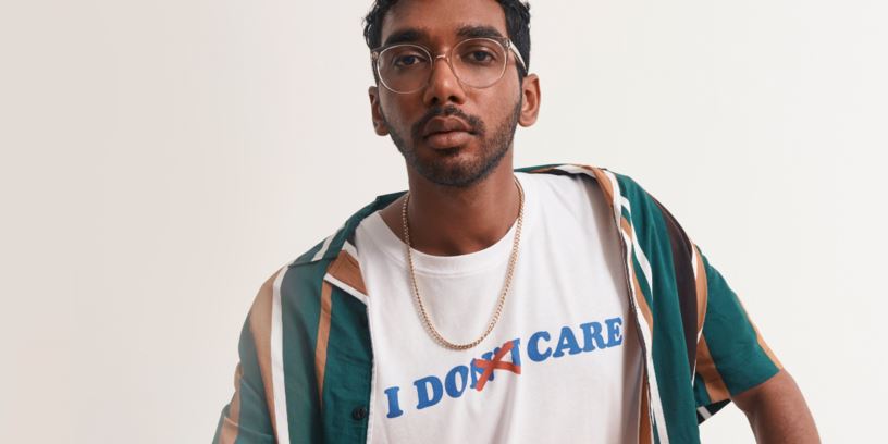 Kille med glasögon och guldkedja runt halsen och en vit t-shirt. På tröjan står texten "I don't care" med överstryken text så att betydelsen blir "I do care". 