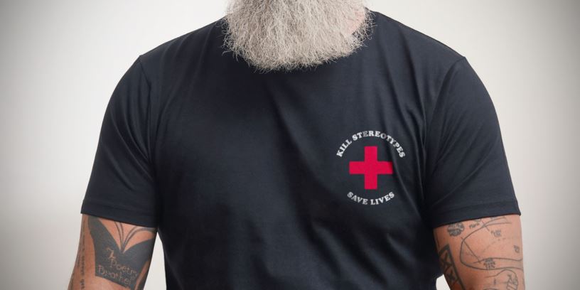 Skäggig man i svart t-shirt och tatuerade armar. På tröjan finns ett tryck med ett rött kors och texten "Kill stereotypes. Save lives". 